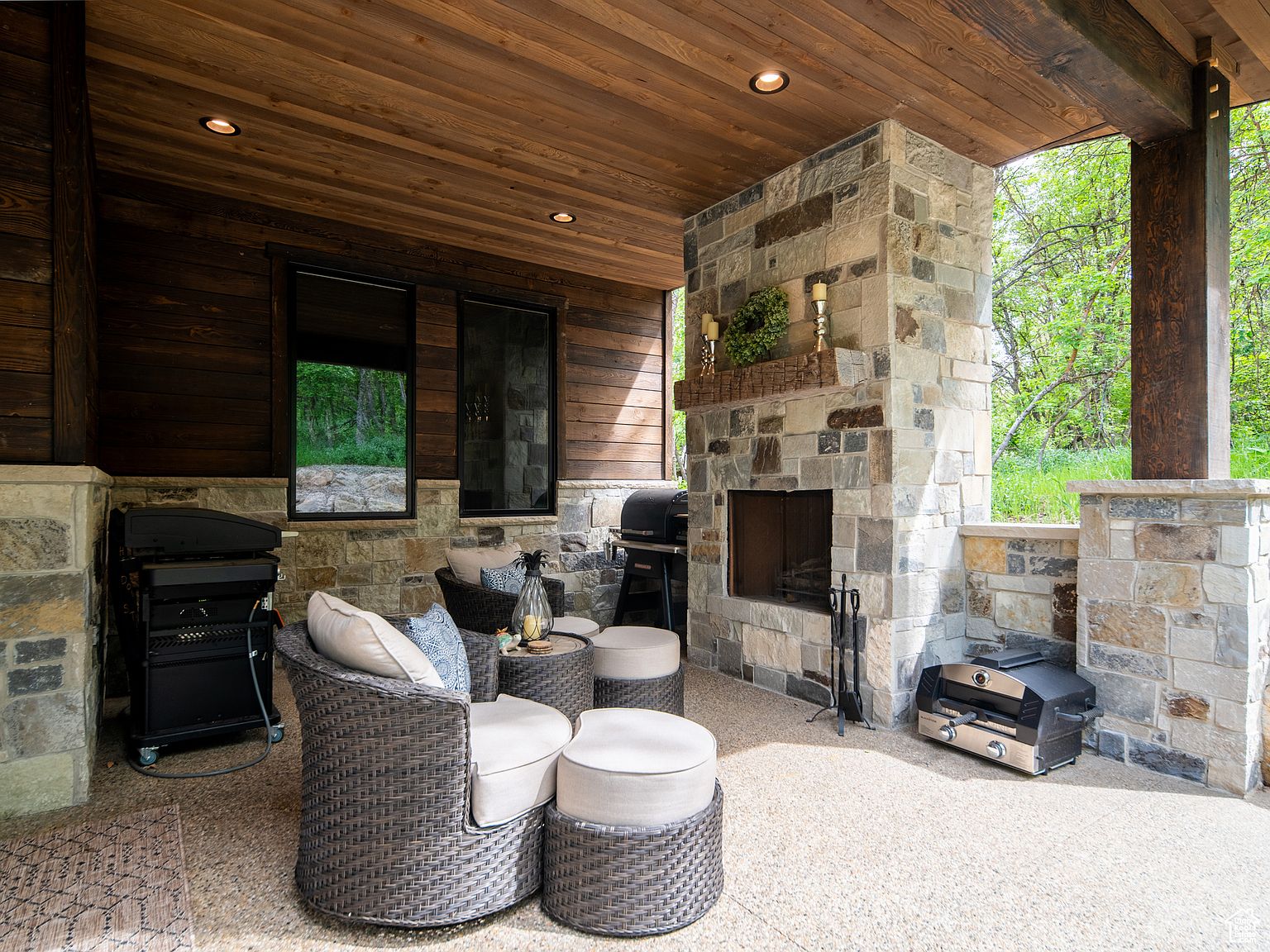 This image showcases a luxurious outdoor patio area, featuring comfortable wicker seating arranged around a stone fireplace. The space includes built-in grilling stations and is covered by a wooden ceiling with recessed lighting. The patio offers a seamless transition between indoor and outdoor living, perfect for entertaining or relaxing.