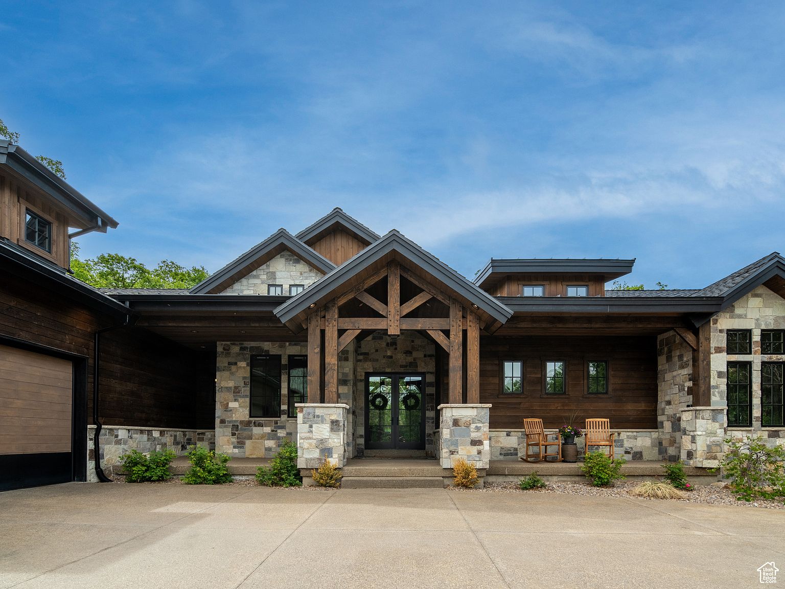 This is a front exterior view of a luxurious, modern home with a rustic touch. The house features a combination of stone and wood siding, a prominent timber-framed entryway, and a well-manicured front yard. The design emphasizes natural materials and a seamless blend with the surrounding landscape, creating an inviting and upscale curb appeal.