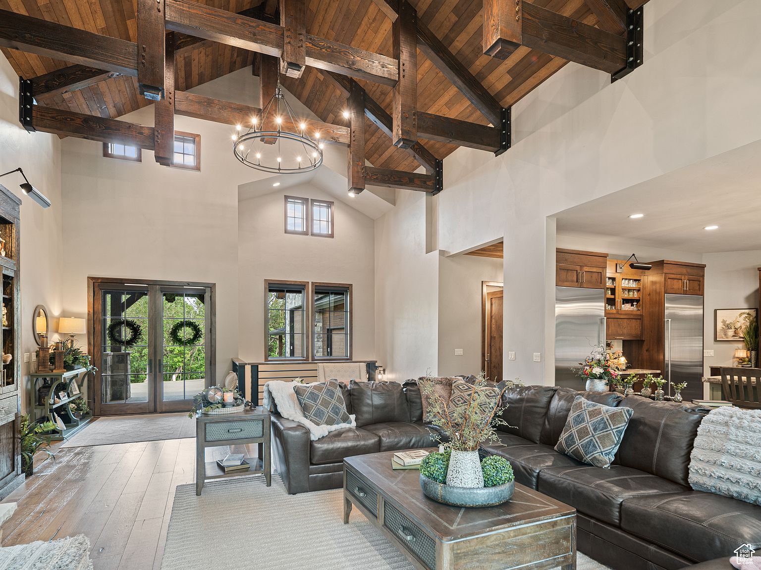 This is an interior shot of a living room featuring a high vaulted ceiling with exposed wooden beams and a large leather sectional sofa. The room is well-lit with natural light coming through multiple windows and a glass door, complemented by a decorative chandelier. The overall impression is one of rustic elegance and spaciousness, ideal for comfortable living and entertaining.