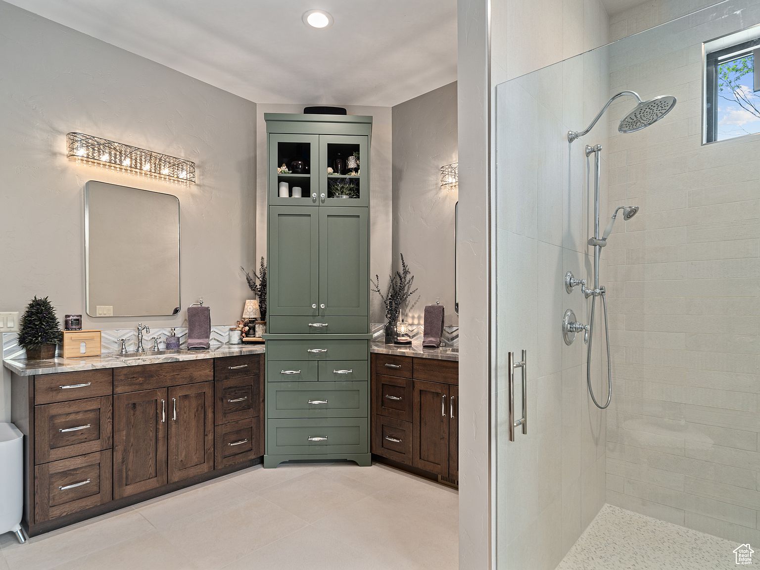 This is a well-lit primary bathroom featuring a double vanity with dark wood cabinets and a light-colored countertop. A tall, green cabinet provides additional storage. The shower is enclosed with glass and has a modern showerhead and fixtures. The overall impression is clean, modern, and luxurious.