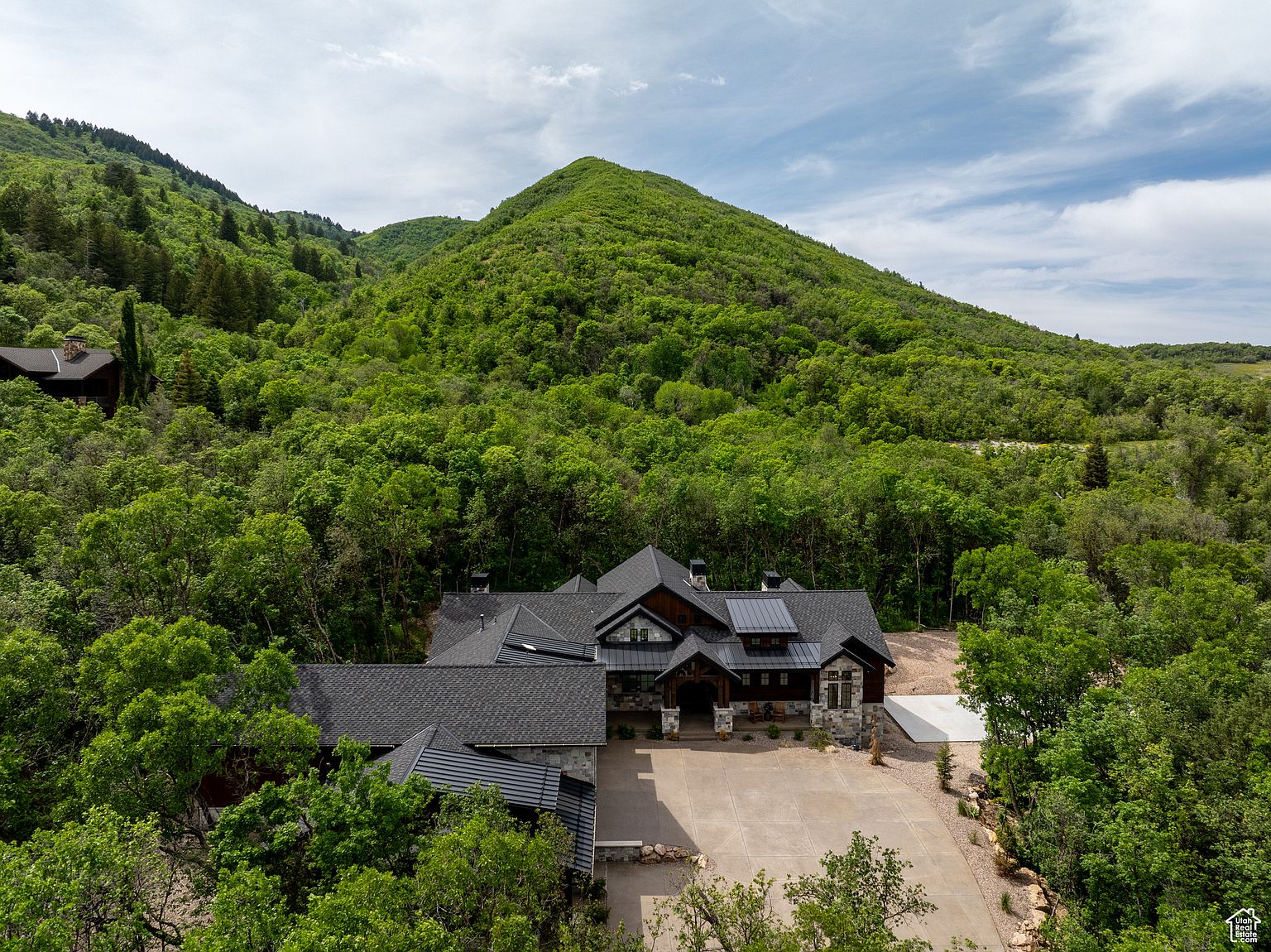 This aerial shot showcases a luxurious home nestled amidst lush greenery and a backdrop of rolling hills. The house features a dark roof, a spacious driveway, and a well-manicured lawn, creating a sense of privacy and tranquility. The architectural design blends seamlessly with the natural surroundings, offering a serene and upscale living environment.