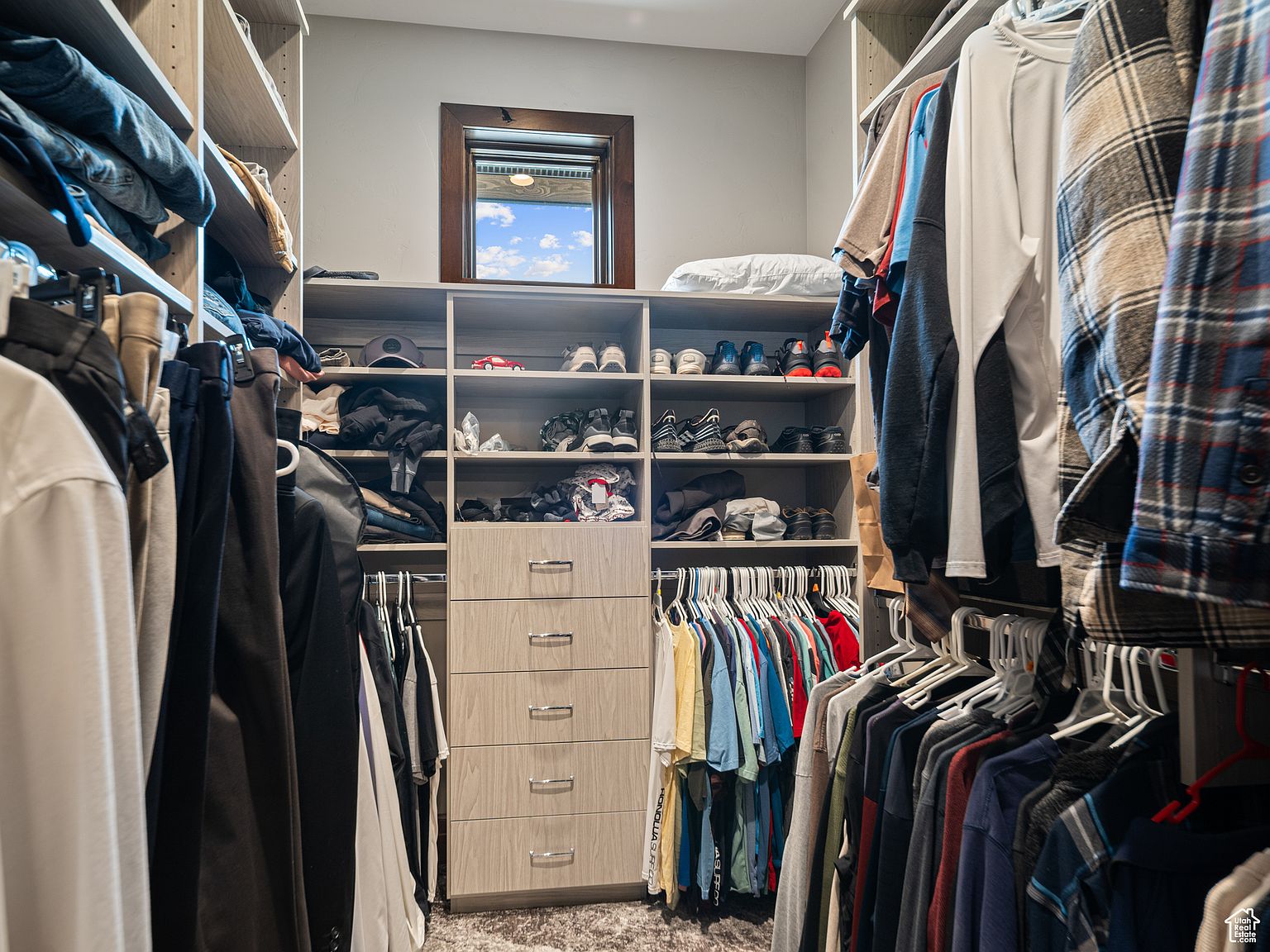 This is a well-organized walk-in closet featuring custom shelving and drawers. Clothes are neatly hung and folded, maximizing space. A small window provides natural light, enhancing the overall appeal and functionality of the closet.