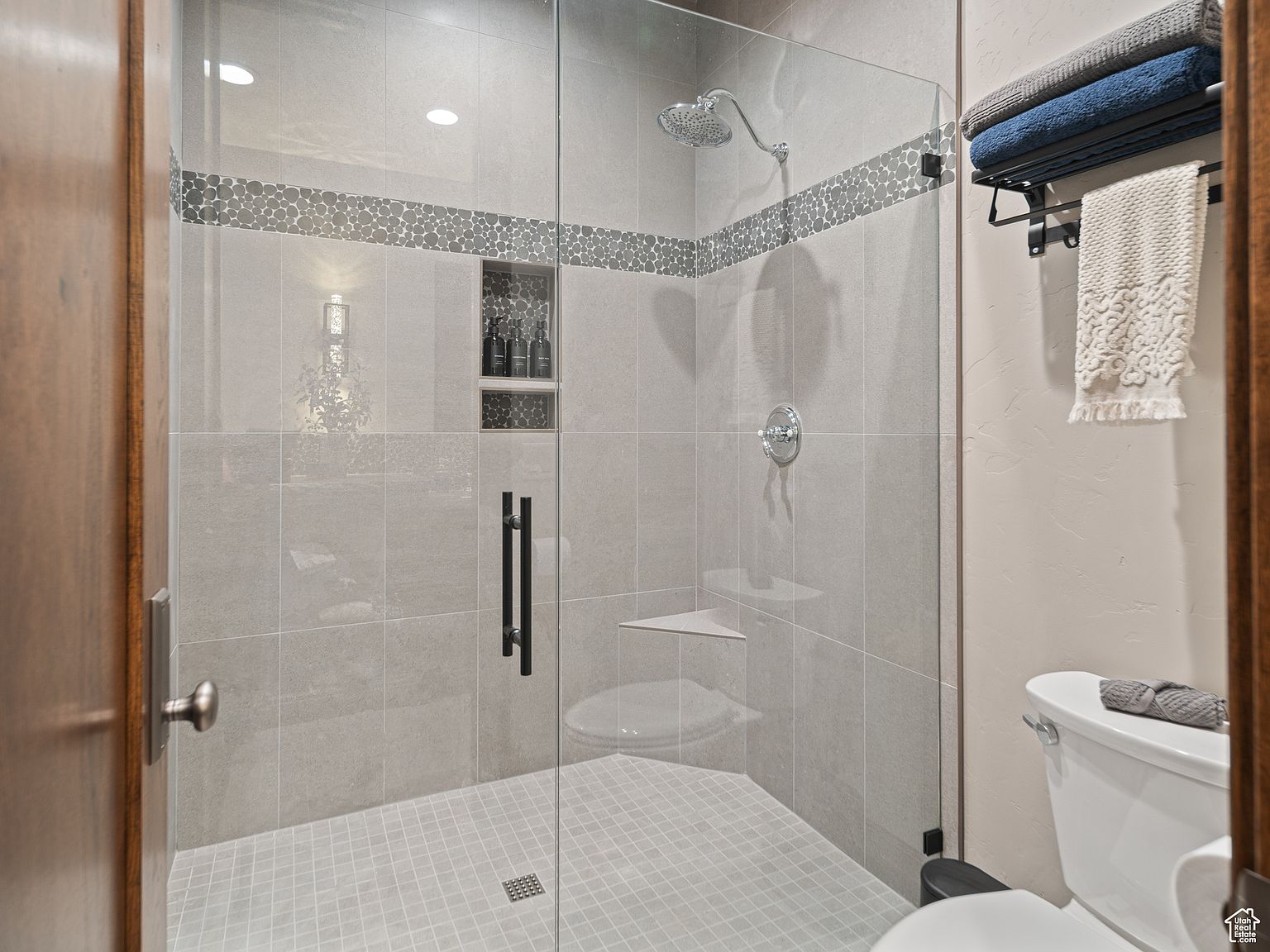 This is a well-lit primary bathroom featuring a modern walk-in shower with glass enclosure, gray tiled walls with a decorative pebble-style accent strip, and a built-in niche for toiletries. A white toilet is visible to the right, and a towel rack with neatly folded towels adds a touch of luxury. The overall impression is clean, contemporary, and spa-like.