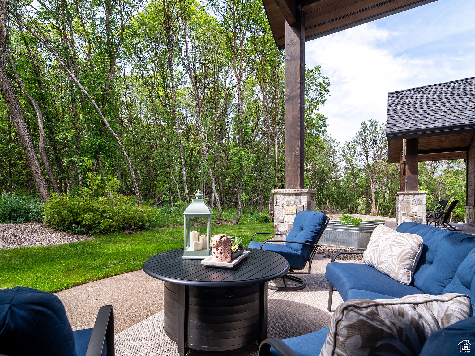 This image showcases a beautifully designed outdoor patio area, featuring comfortable blue cushioned seating arranged around a modern black fire pit table. The patio is surrounded by lush greenery and mature trees, creating a serene and private atmosphere. The space is partially covered by a wooden structure with stone pillars, offering shade and architectural interest, making it an ideal spot for relaxation and entertaining.