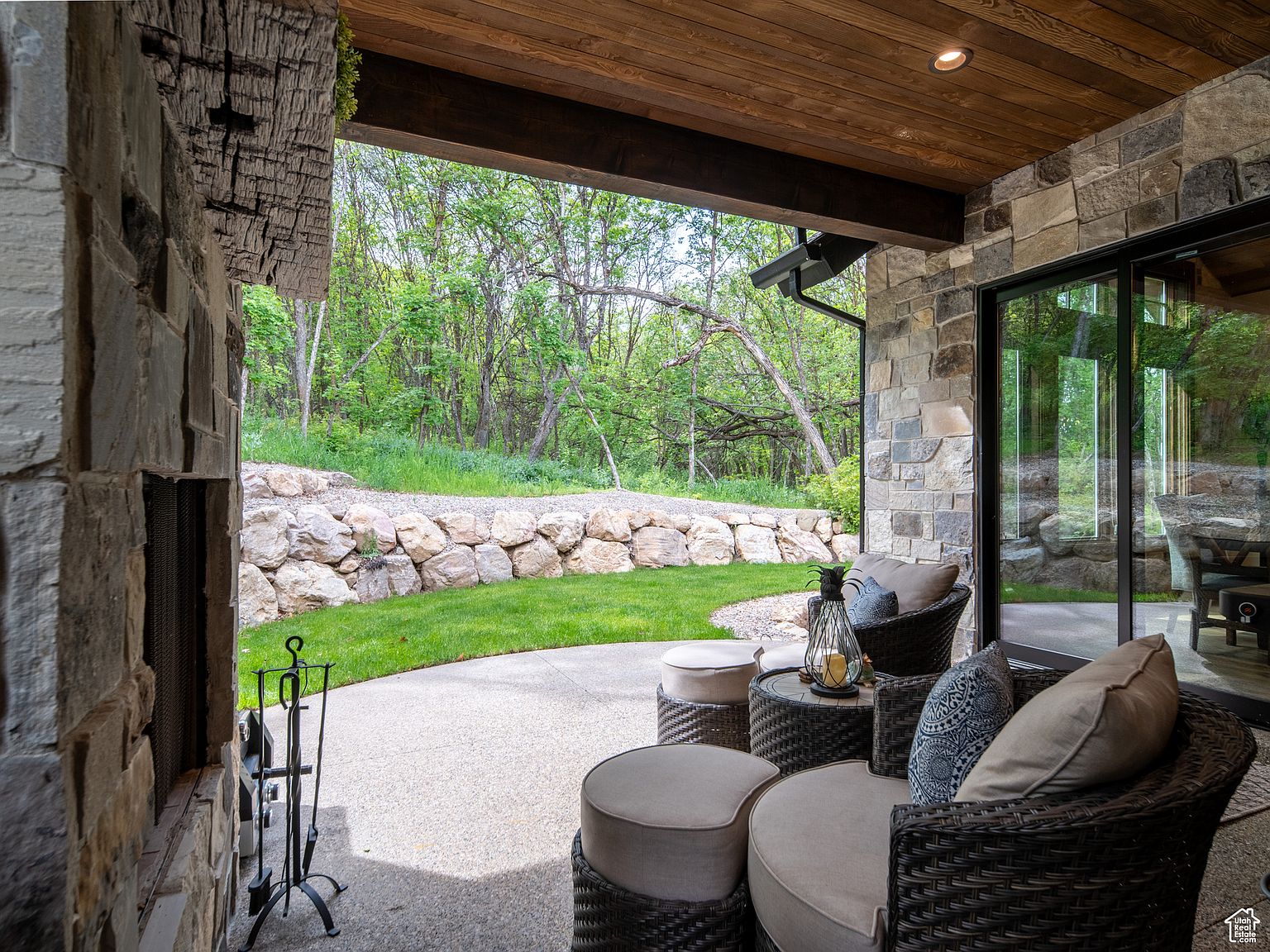 This image showcases a luxurious outdoor patio area, featuring comfortable wicker seating with plush cushions arranged around a small table. The patio is constructed with a smooth concrete surface and is partially covered by a wooden ceiling, providing shade. A stone wall and lush greenery create a serene backdrop, enhancing the property's appeal for relaxation and outdoor entertainment.