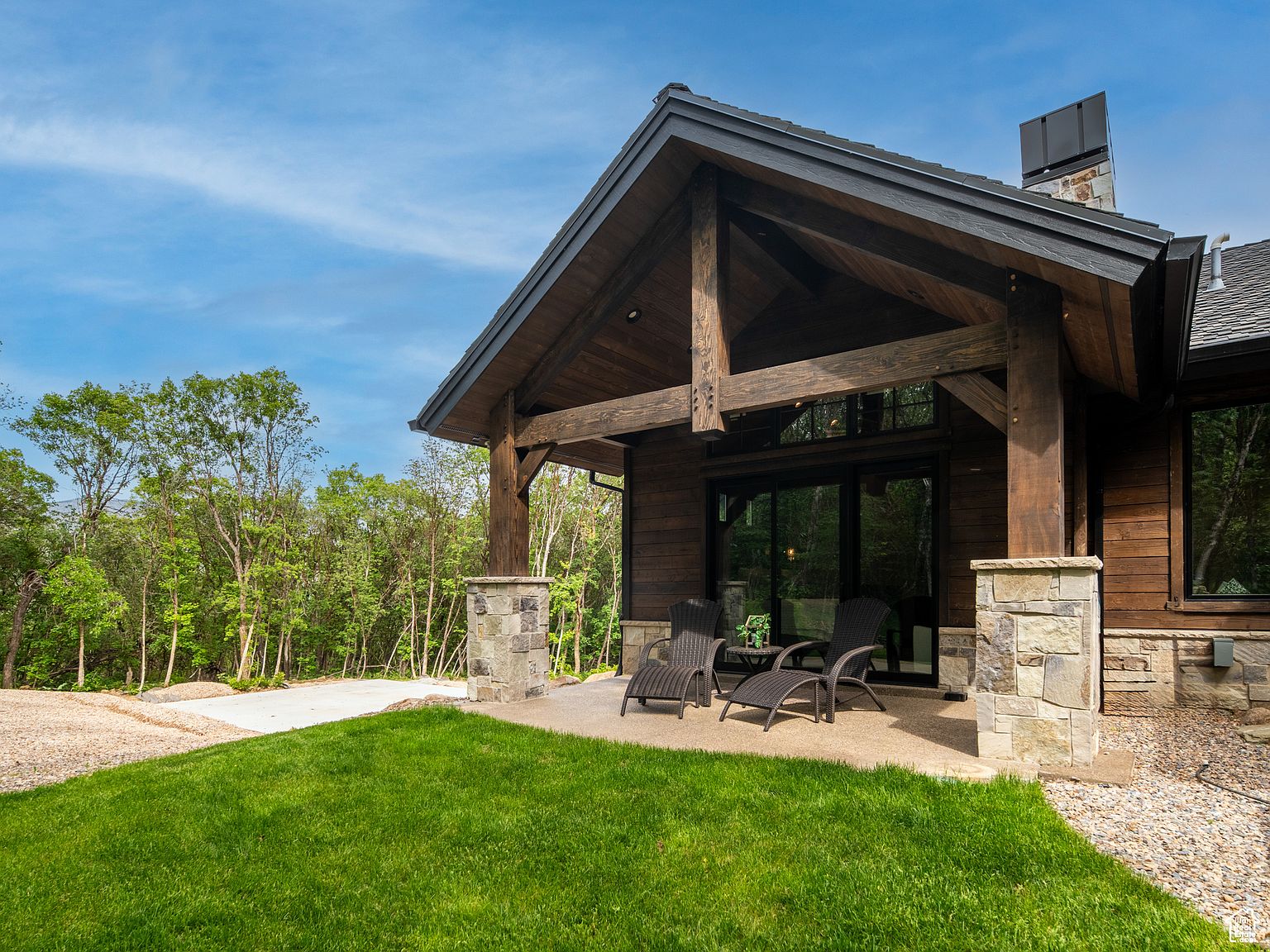 This image showcases a beautifully designed patio area attached to a rustic-style home. The patio features stone pillars supporting a timber-framed roof, creating a cozy outdoor living space. Wicker lounge chairs are arranged on the concrete patio, inviting relaxation, while the surrounding green lawn and trees provide a serene backdrop, enhancing the property's appeal.
