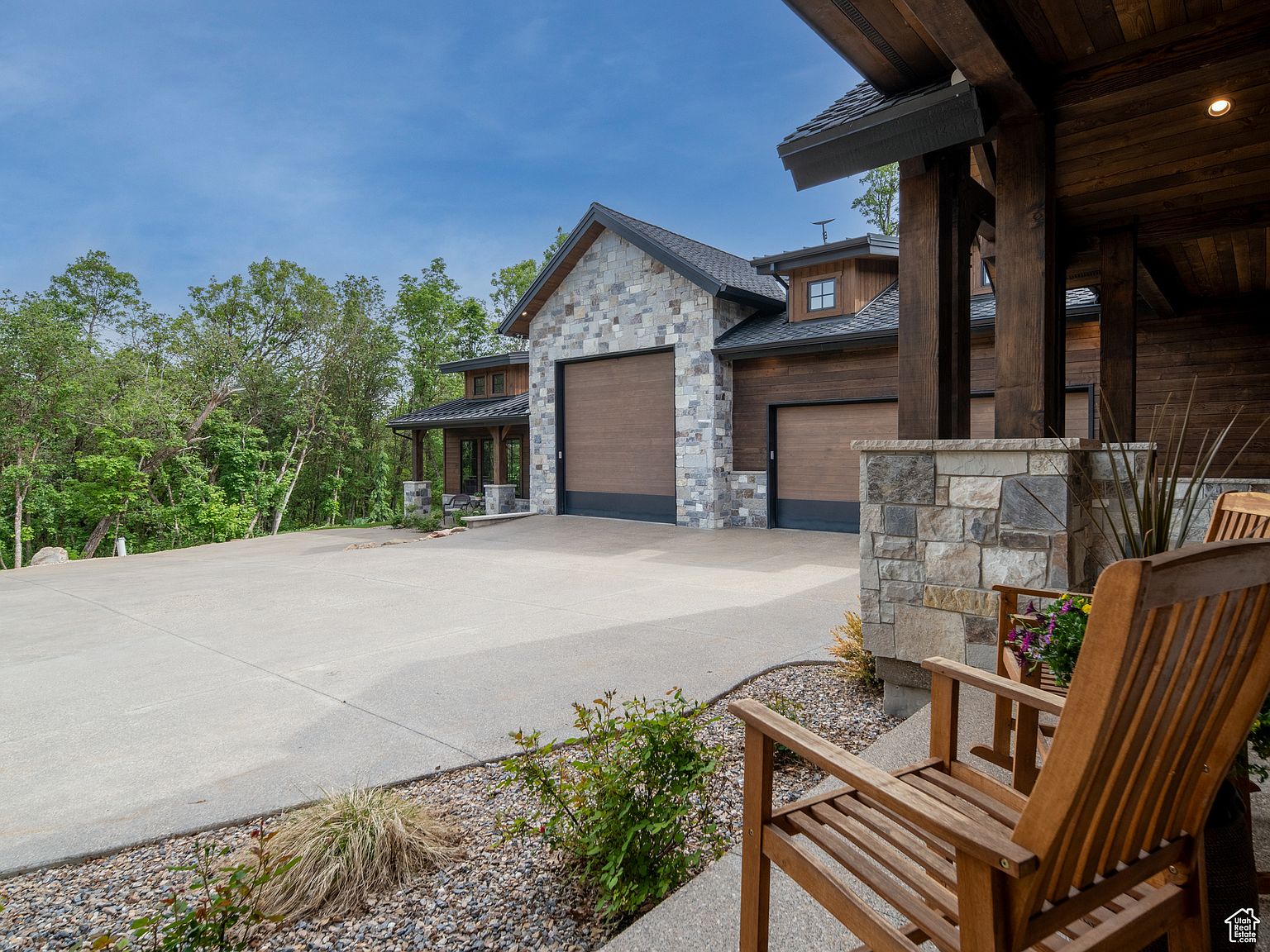 This is a front exterior view of a luxurious home featuring a stone and wood facade. The house has a large driveway leading to a multi-car garage with wooden doors. The landscaping includes gravel beds with shrubs and a wooden porch with chairs, creating a welcoming and high-end impression.