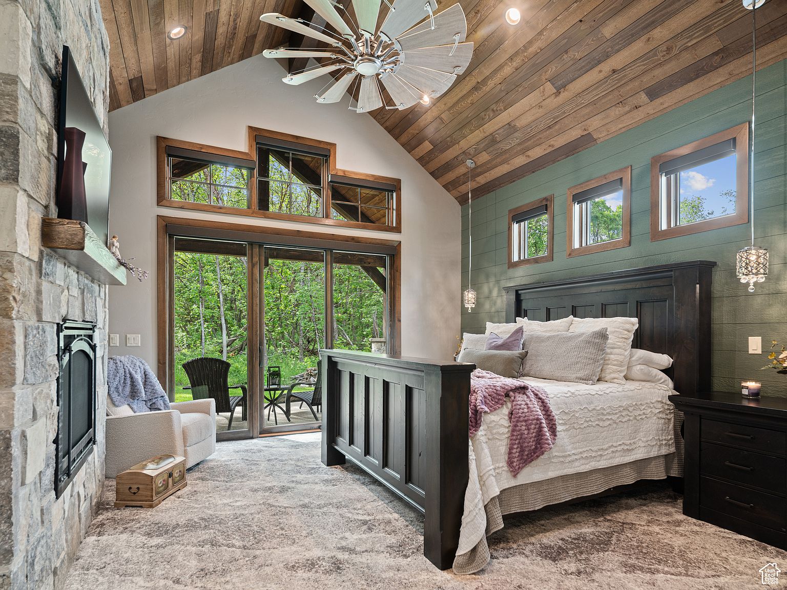 This is a primary bedroom featuring a large bed with a dark wood frame, complemented by a stone fireplace and a wall of windows overlooking a lush green landscape. The room has a rustic-chic aesthetic with a wood-paneled ceiling and a mix of textures, creating a cozy and luxurious atmosphere. The perspective is from the foot of the bed, showcasing the entire room.
