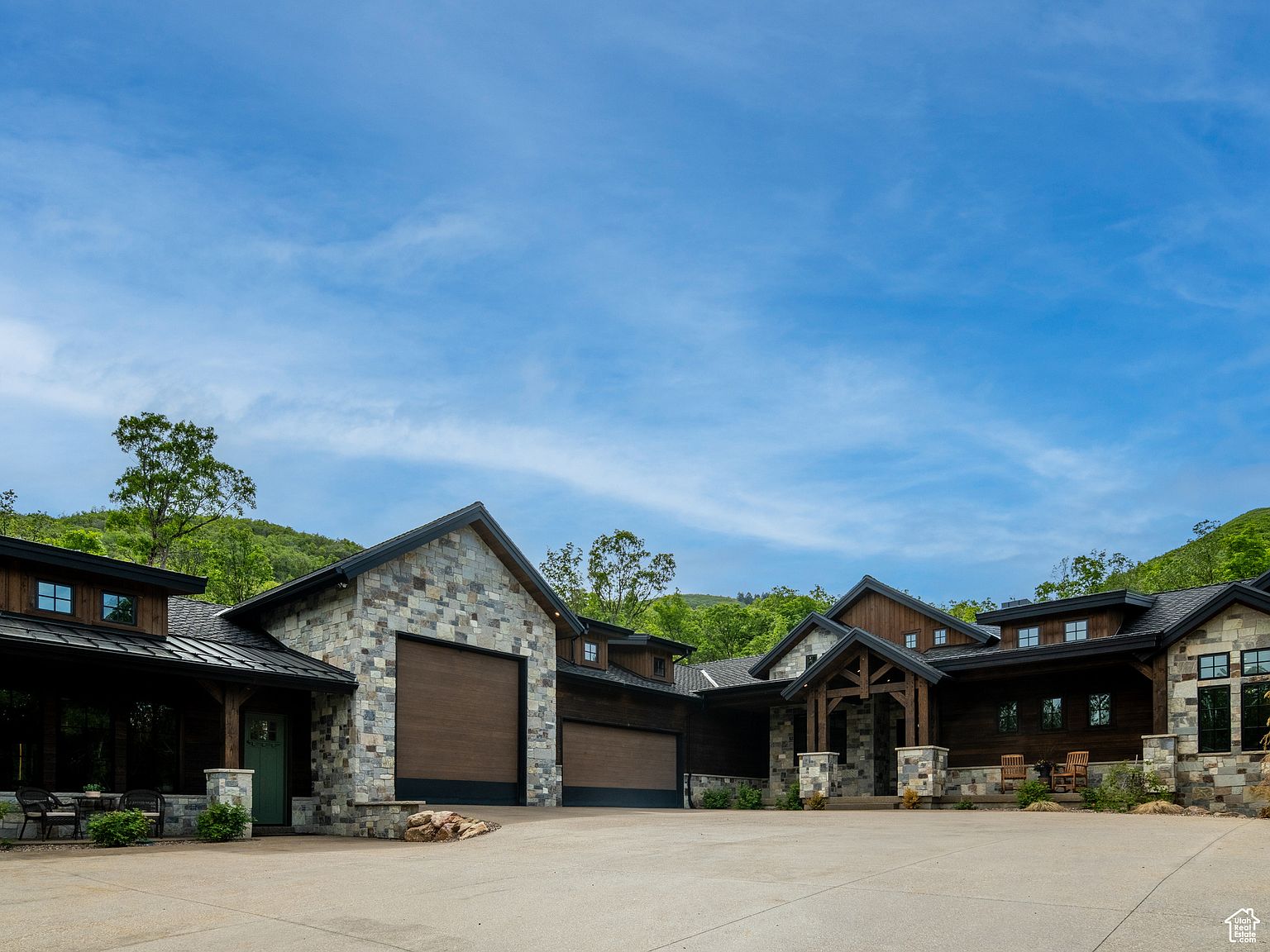 This is a front exterior view of a large, luxurious home with a stone and wood facade. The house features multiple garage doors, a covered entryway with stone pillars, and a well-maintained concrete driveway. The surrounding landscape includes lush greenery and a clear blue sky, creating an inviting and impressive curb appeal.