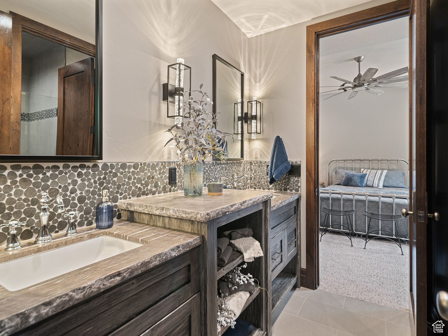 This is a well-lit bathroom featuring a double vanity with a stone countertop and dark wood cabinetry. The backsplash is decorated with pebble-style tiles, and modern sconces flank the mirrors. An open doorway reveals a glimpse into an adjacent bedroom, creating a sense of flow and connection between the spaces.