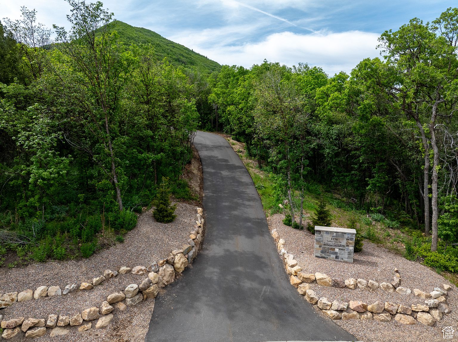 This image showcases the front view of a property, emphasizing the long, paved driveway leading up to the house. The driveway is bordered by meticulously arranged rock landscaping and lush greenery, creating a sense of privacy and natural beauty. A stone sign marks the entrance, adding a touch of elegance and clearly displaying the property's address.