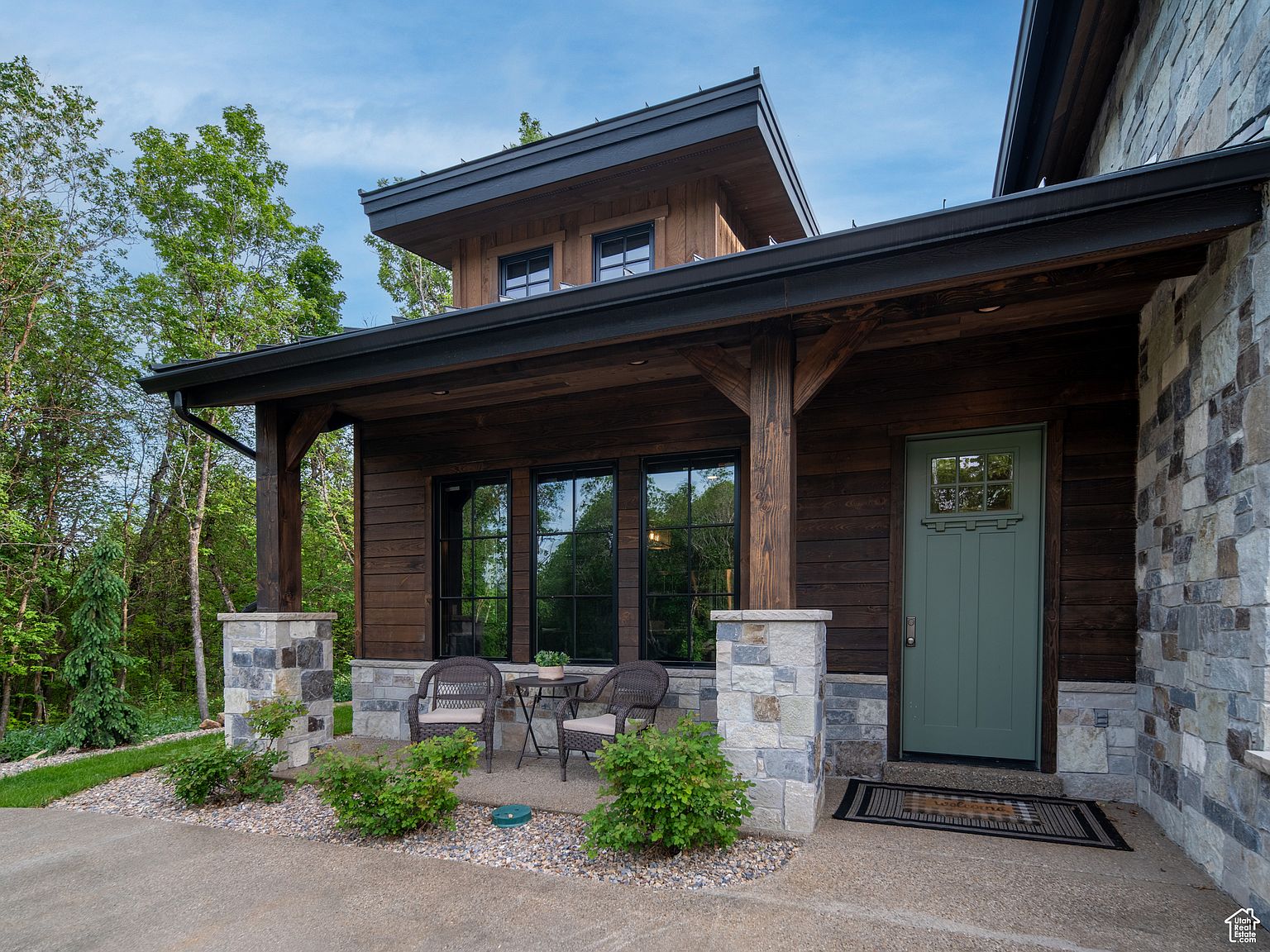 This image showcases the entryway of a home, featuring a sage green front door flanked by stone pillars and dark wood siding. A small seating area with wicker chairs and a table is visible on the porch. The overall impression is one of rustic elegance and inviting curb appeal.