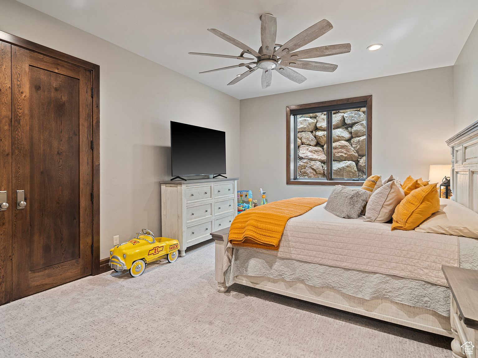 This is a cozy bedroom featuring a bed with a cream-colored comforter and yellow accent pillows. A dresser with a TV on top sits against the wall, and a small window provides a view of a rock wall outside. A wooden door and a vintage yellow toy car add character to the room.