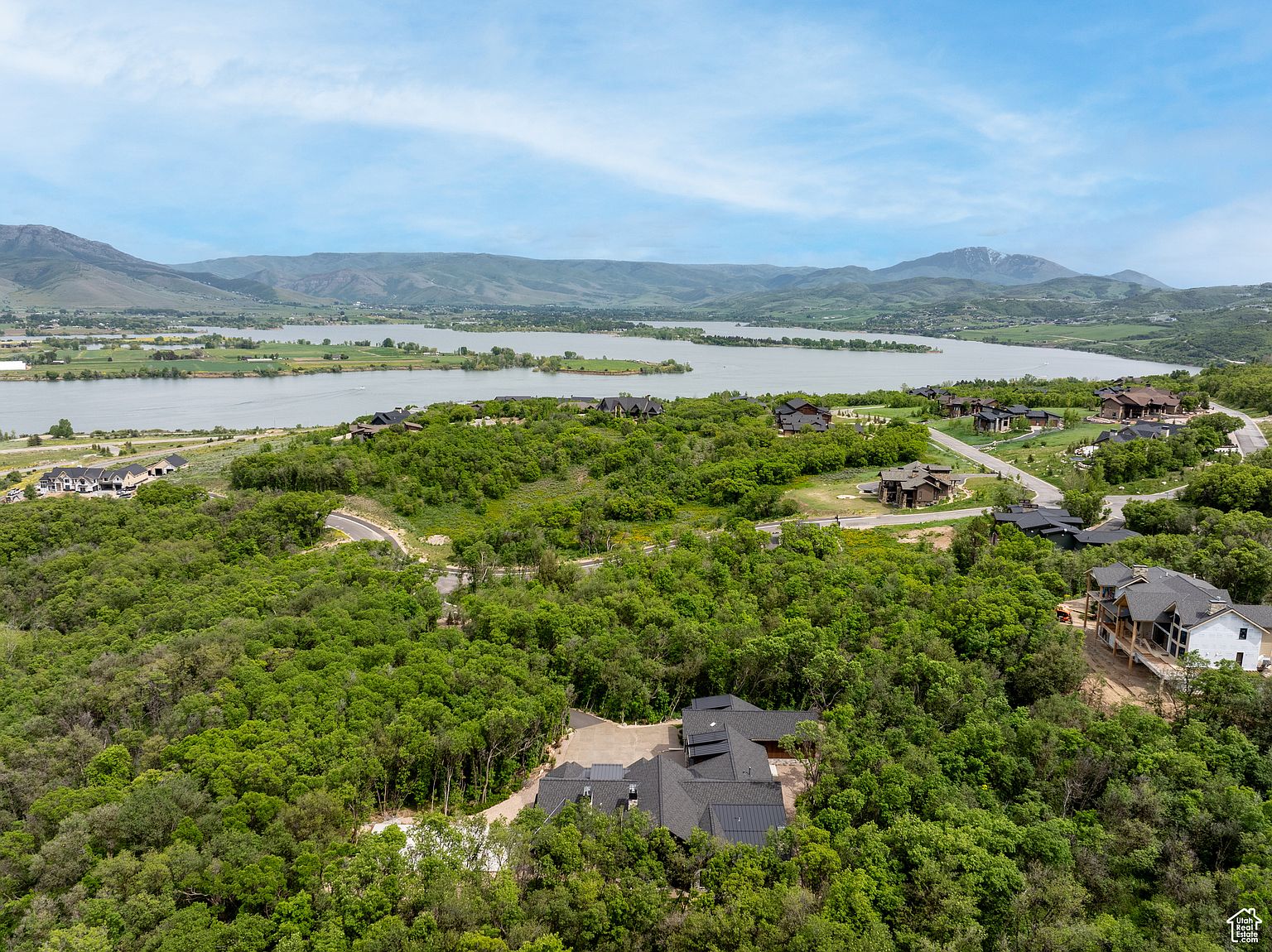This aerial view showcases a luxurious home nestled amidst lush greenery, offering privacy and stunning natural surroundings. The property features a dark roof, a spacious driveway, and is surrounded by mature trees. In the background, a serene lake and rolling hills create a picturesque backdrop, enhancing the property's appeal and highlighting its desirable location.