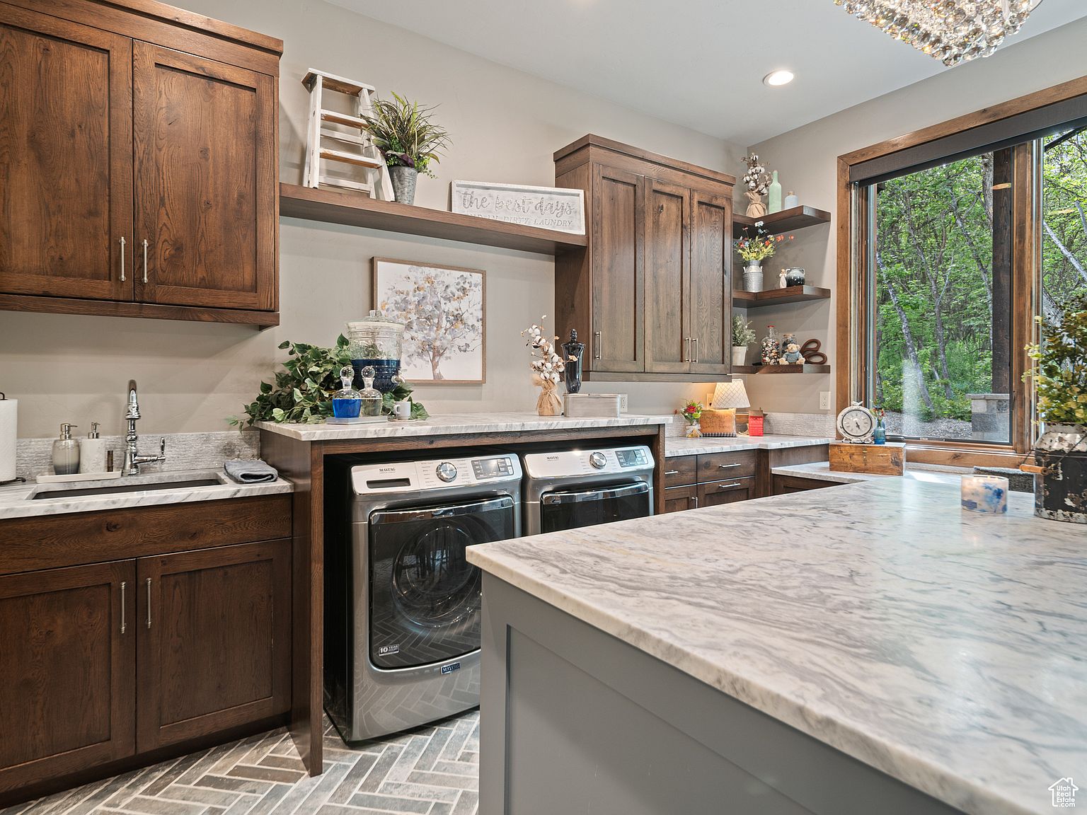 This is an interior shot of a laundry room featuring dark wood cabinetry, marble countertops, and modern appliances. A front-loading washer and dryer are visible, along with decorative shelving and a window providing natural light. The room has a clean and organized feel, suggesting a functional and stylish space.