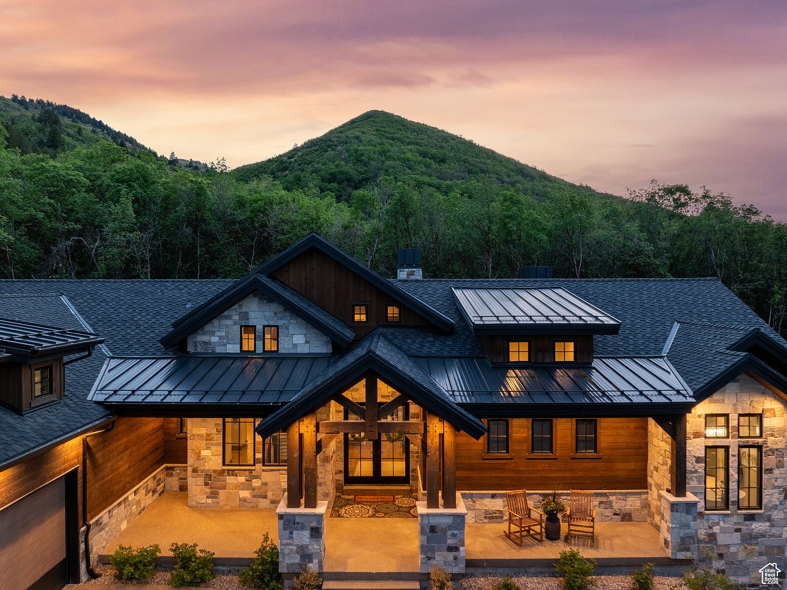 This is a front exterior view of a luxurious mountain home at dusk. The house features a combination of stone and wood siding, a dark roof, and a welcoming entryway with wooden pillars. The backdrop includes a lush green mountain and a colorful sunset sky, enhancing the property's appeal and highlighting its serene setting.