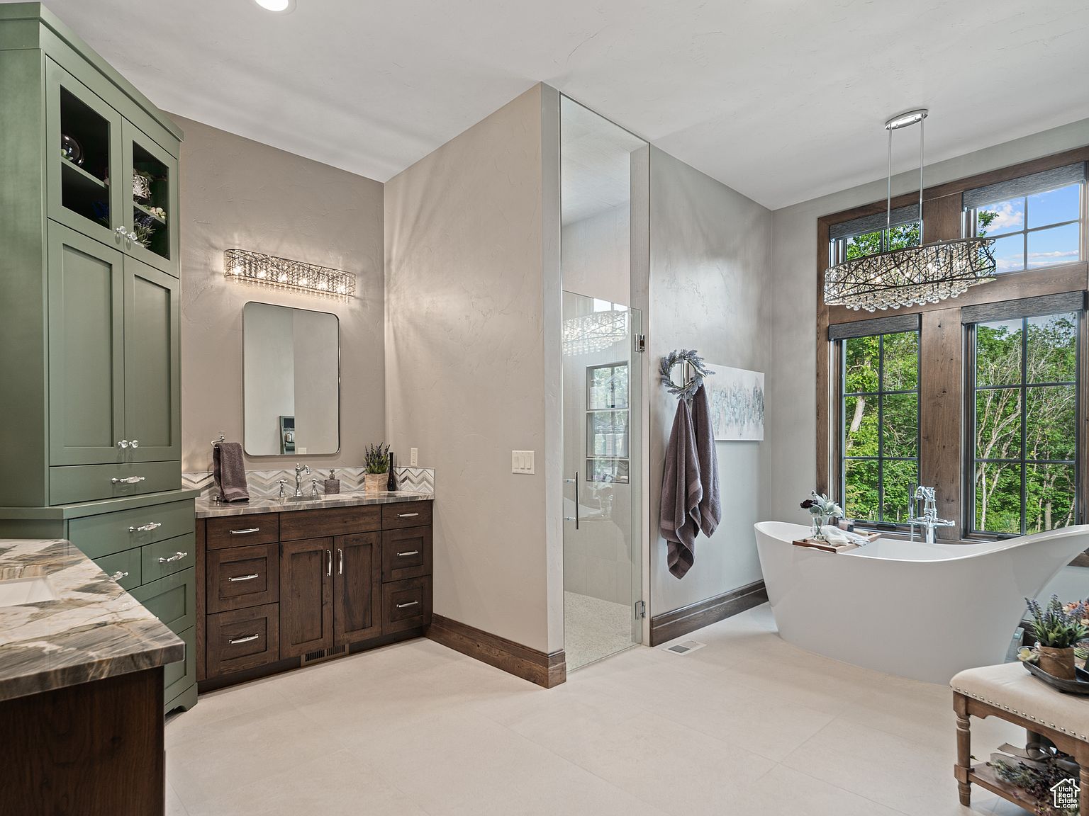 This is a luxurious primary bathroom featuring a freestanding soaking tub positioned beneath a large window offering natural light and a view of the outdoors. The bathroom includes a glass-enclosed shower, a dark wood vanity with double sinks, and a tall green storage cabinet. The overall design is elegant and spa-like, creating a relaxing atmosphere.