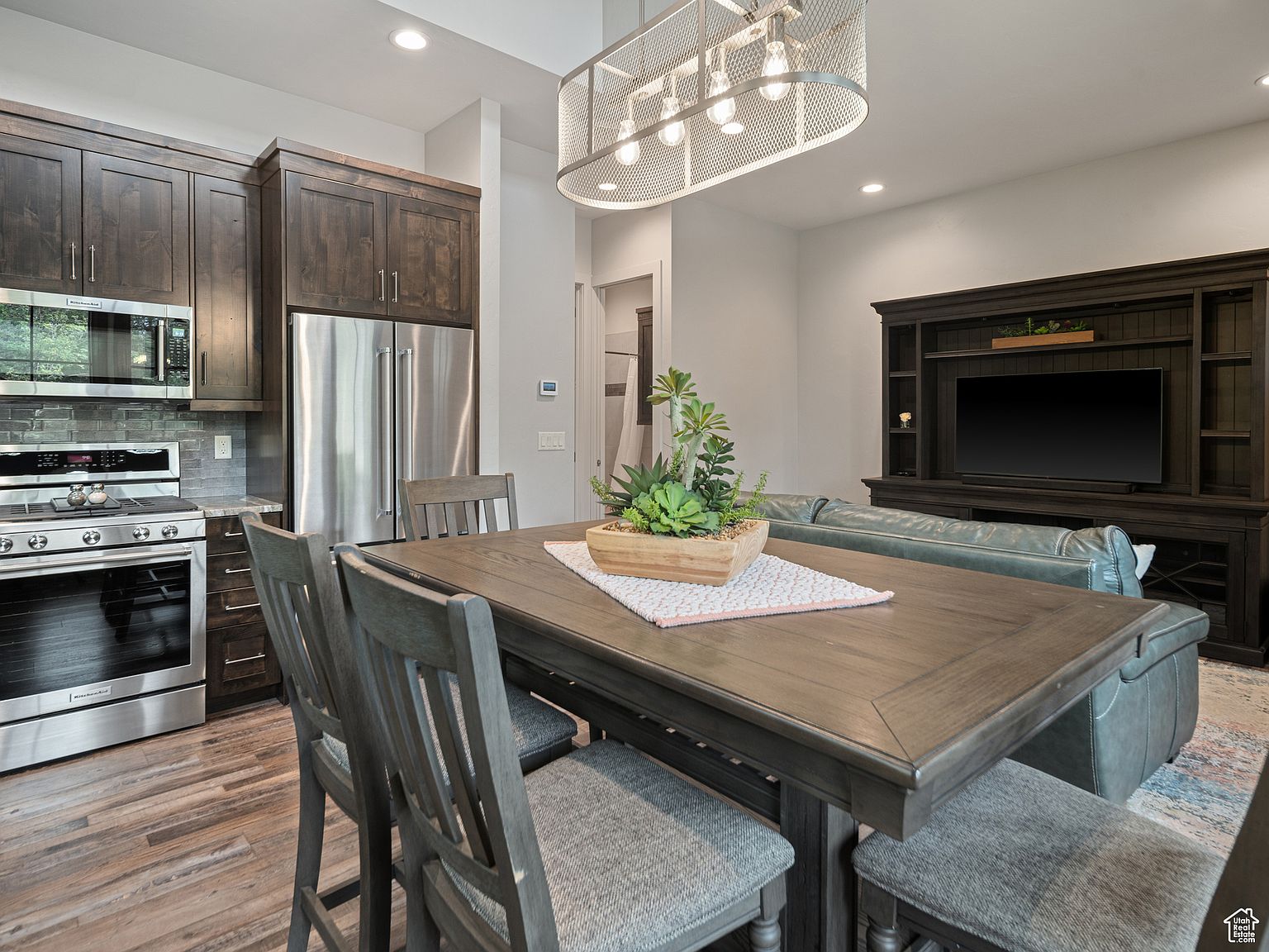 This interior shot showcases a dining area adjacent to the kitchen and living room. The dining table, crafted from dark wood, is the focal point, adorned with a centerpiece of succulents. The kitchen features stainless steel appliances and dark wood cabinetry, while the living room boasts a large entertainment center and a comfortable leather sofa, creating a cohesive and inviting living space.