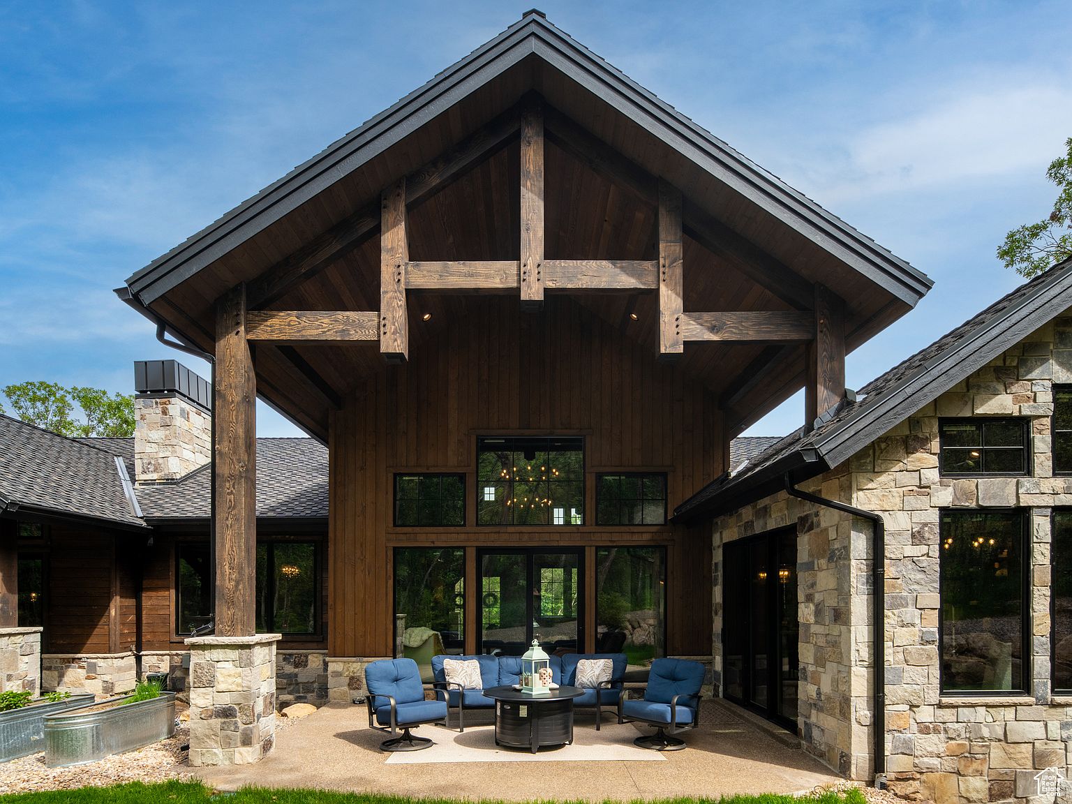 This image showcases a beautifully designed outdoor patio area of a luxury home. The patio features comfortable blue seating around a fire pit, set against the backdrop of a modern rustic home with wood and stone exterior elements. The architectural design includes large windows and a striking roofline, creating an inviting and stylish outdoor living space.