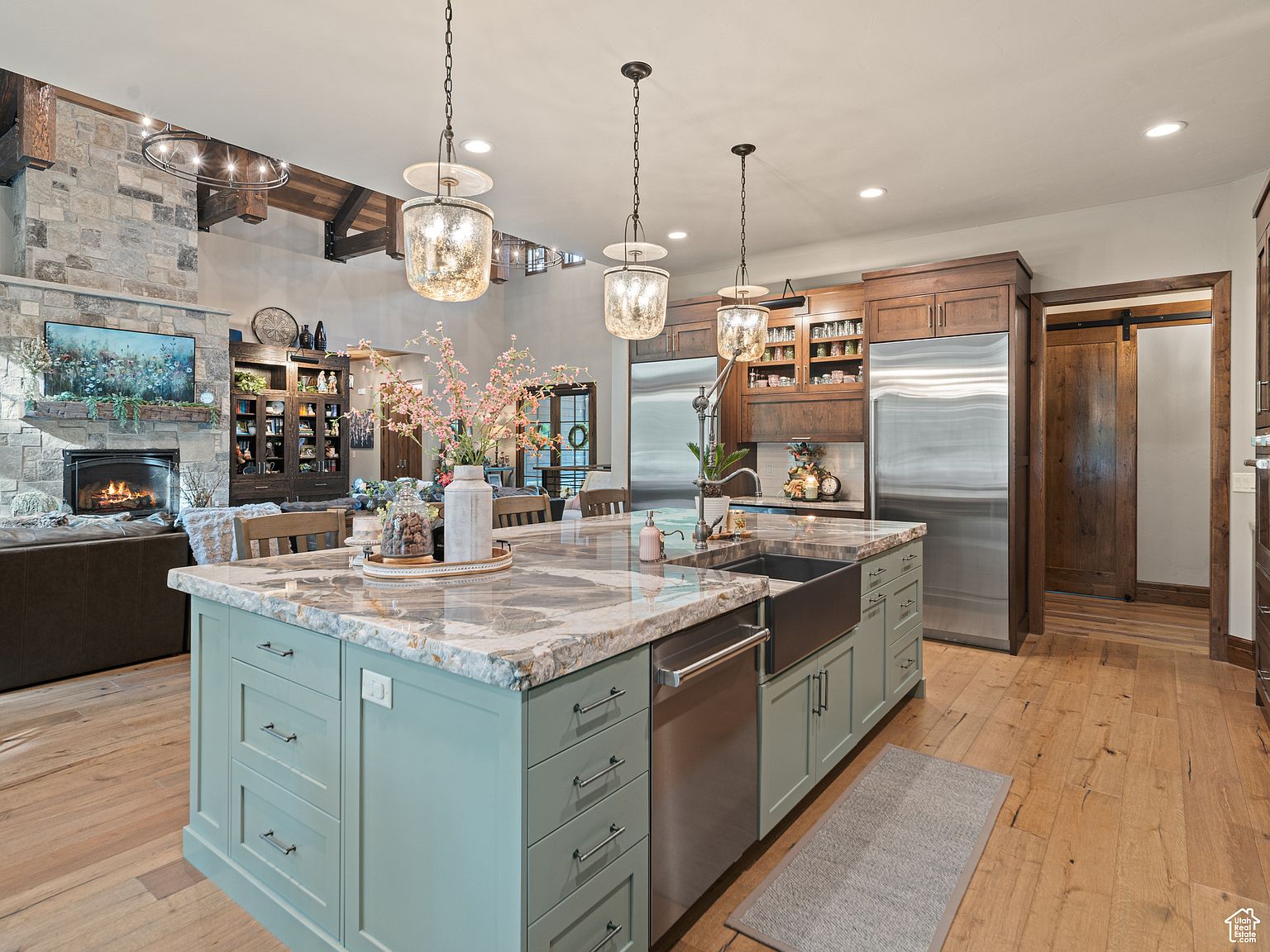 This is a spacious, open-concept kitchen featuring a large island with a marble countertop and light green cabinetry. Stainless steel appliances, including a refrigerator and dishwasher, are integrated into the dark wood cabinetry along the back wall. The kitchen flows seamlessly into a living area with a stone fireplace, creating a warm and inviting atmosphere.