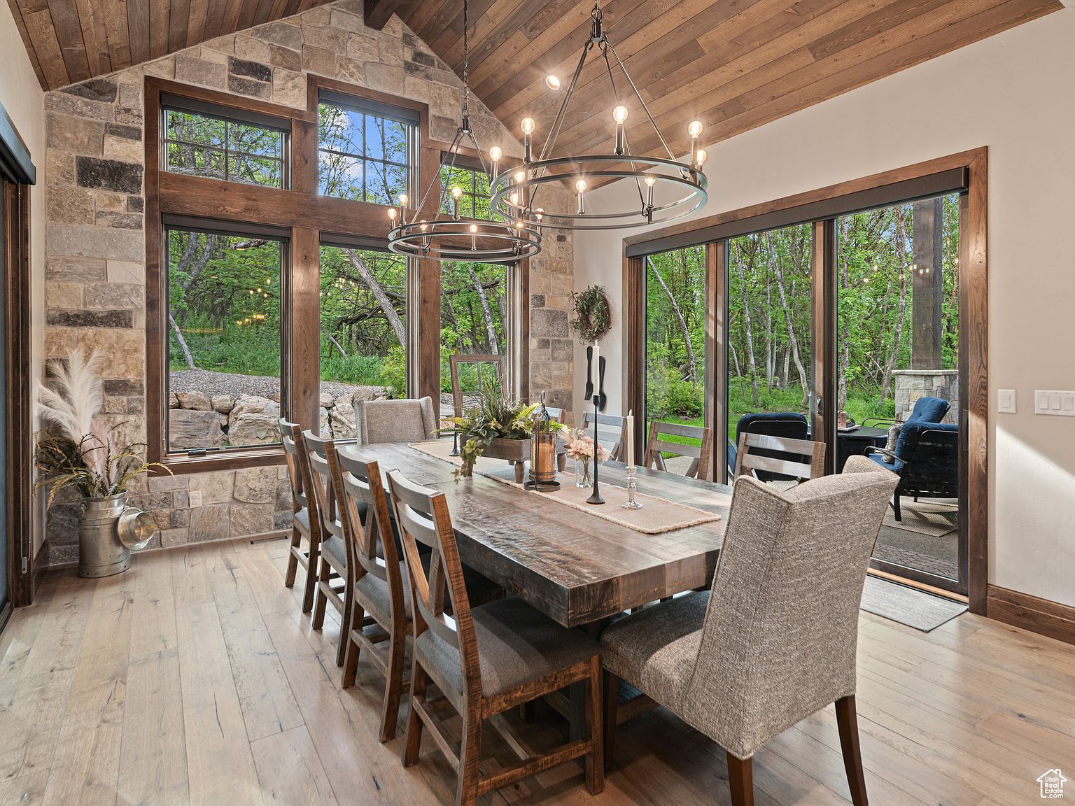 This is an interior shot of a dining room featuring a large wooden table with seating for ten. The room has a rustic yet elegant feel, with stone walls, wooden floors and ceiling, and large windows offering views of the outdoors. Two modern chandeliers hang above the table, providing ample lighting.