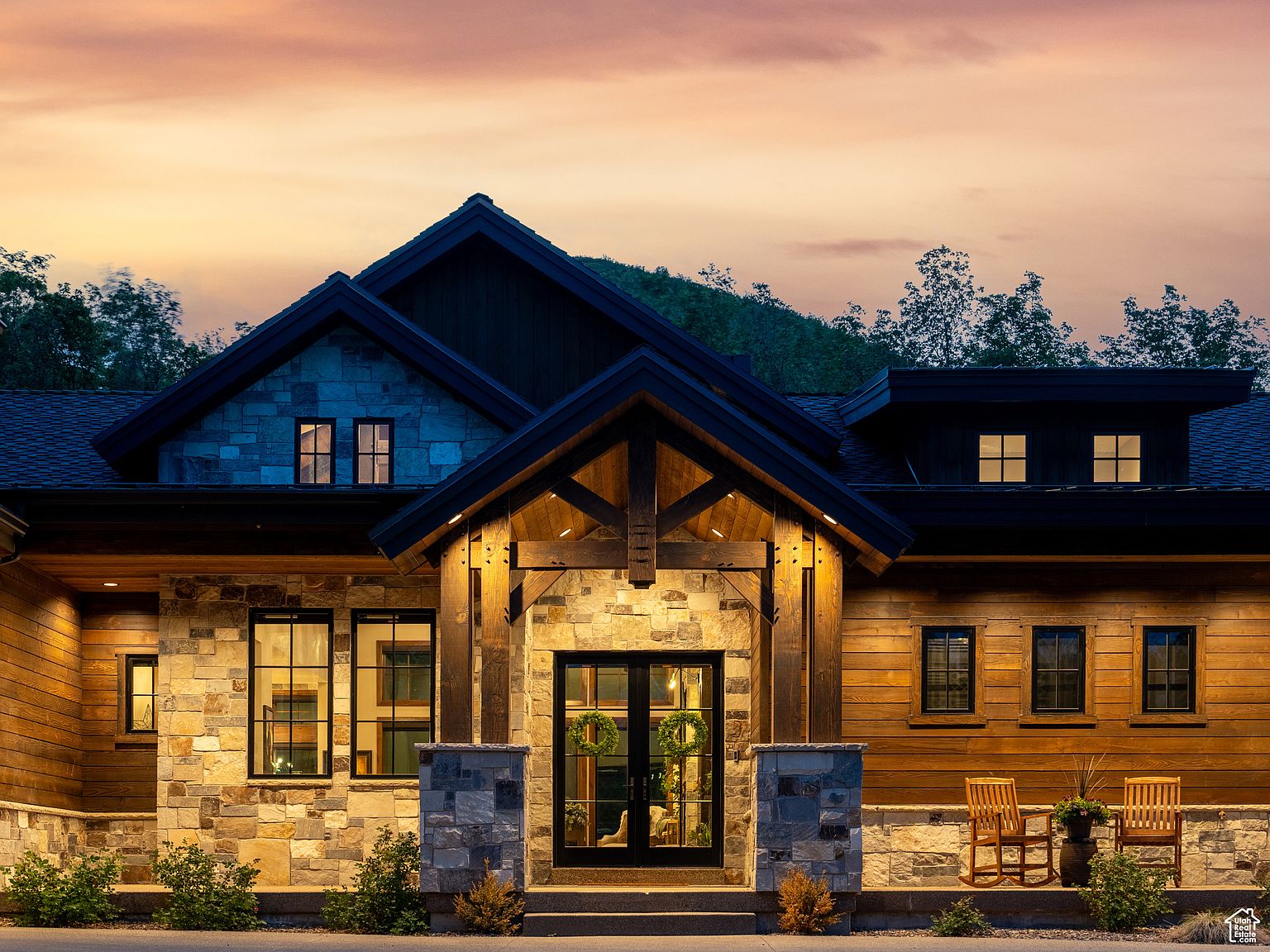 This image showcases the grand entryway of a luxurious home, featuring a combination of stone and wood exterior finishes. The entrance is framed by stone pillars and a wooden gable, with a black front door adorned with wreaths. Two wooden rocking chairs sit on the porch, adding a touch of rustic charm to the elegant facade.