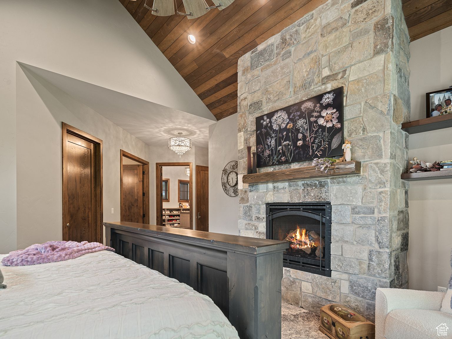 This is a primary bedroom featuring a stone fireplace with a mounted television above the mantle. The room has a vaulted ceiling with wood paneling and includes a view into a hallway with a decorative chandelier. The bed is partially visible in the foreground, and there are doorways leading to other rooms, possibly closets or bathrooms.