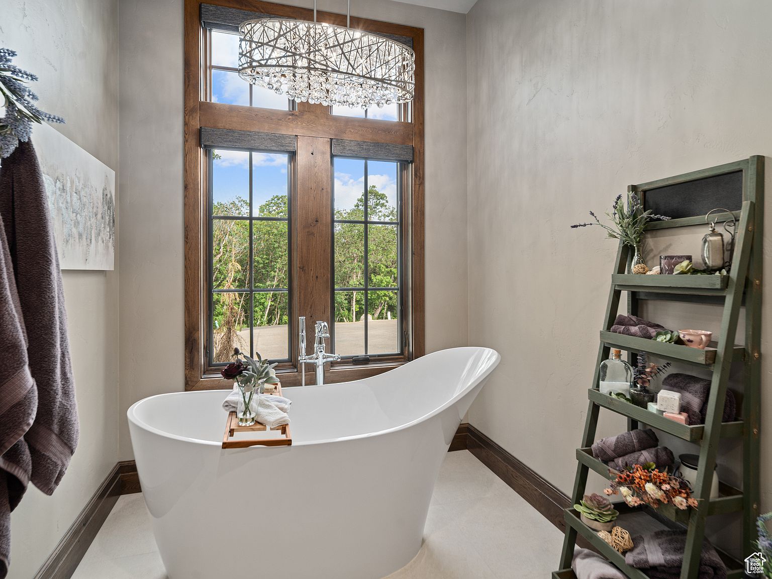 This is a luxurious primary bathroom featuring a freestanding white bathtub positioned in front of a large window that offers a view of lush greenery. A decorative ladder shelf holds towels and bath accessories, adding a touch of rustic charm to the space. The room is illuminated by an elegant chandelier, creating a serene and spa-like atmosphere.