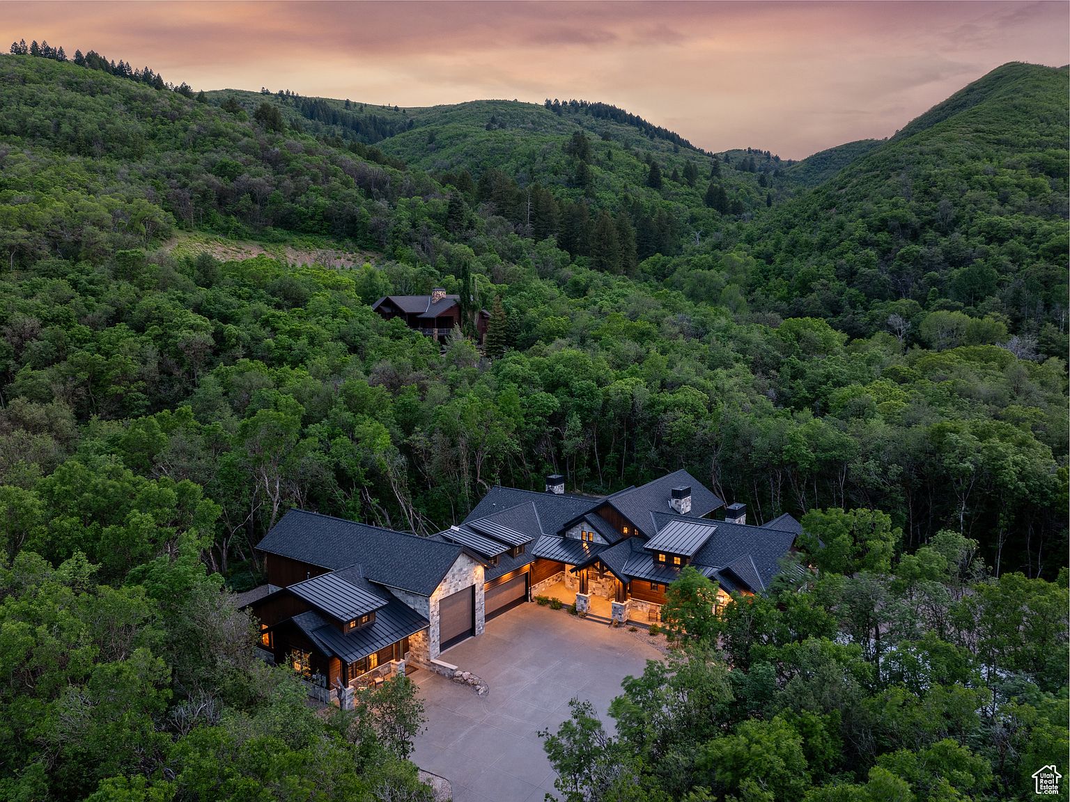 This aerial view showcases a luxurious home nestled in a lush, green mountain setting. The house features a dark roof, stone accents, and a spacious driveway, blending seamlessly with the natural surroundings. The overall impression is one of secluded elegance and privacy.