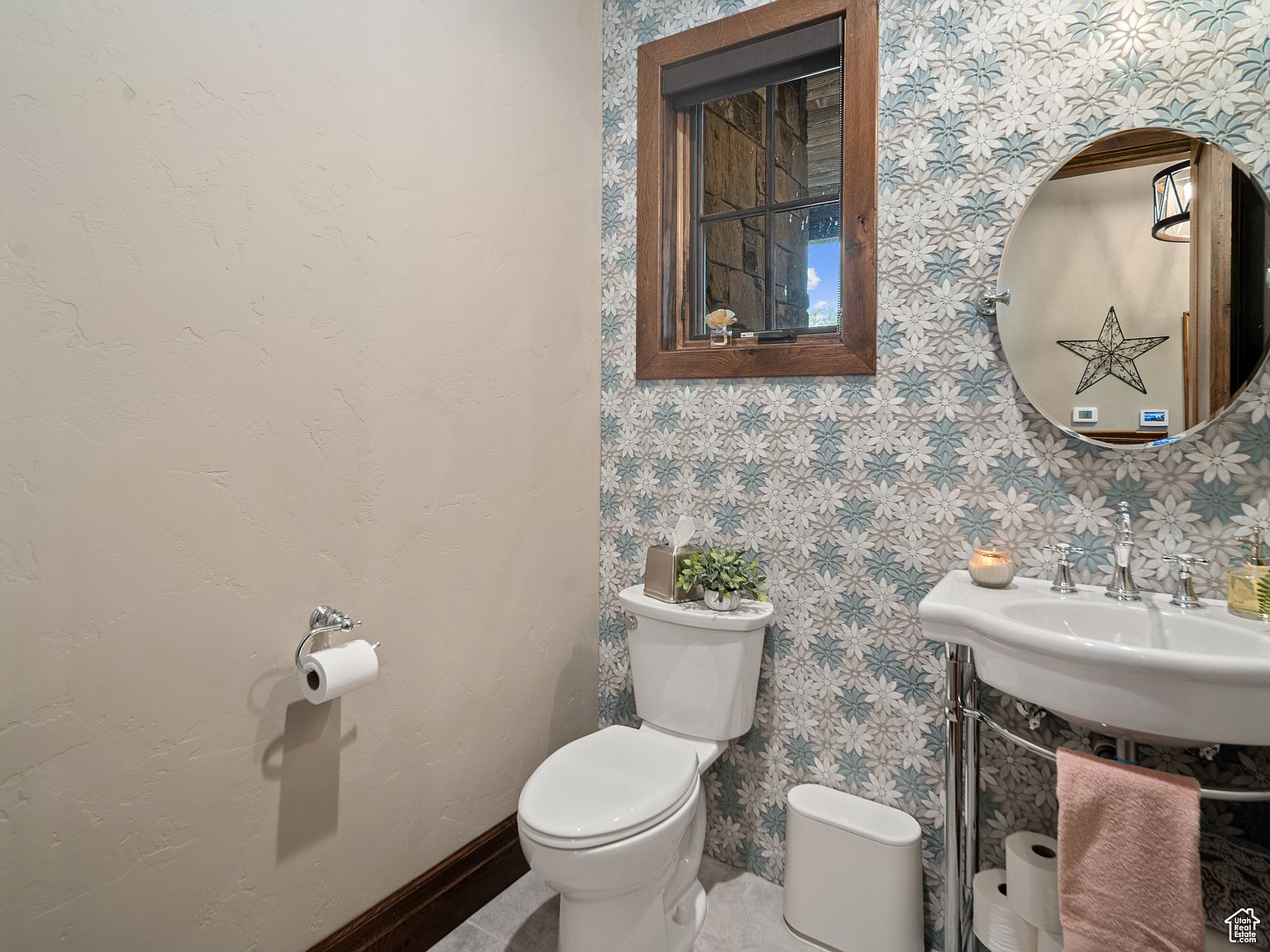 This is a well-decorated guest bathroom featuring a toilet, a pedestal sink, and a window. The walls are partially covered in a floral patterned wallpaper, adding a touch of elegance. The room is clean and well-lit, creating a welcoming atmosphere.