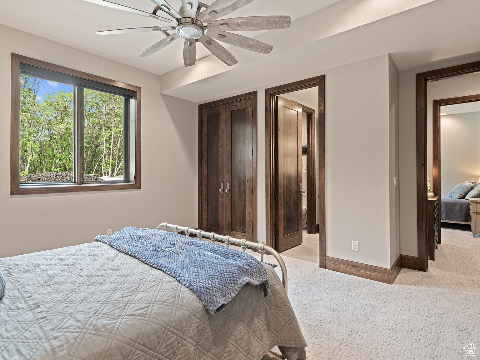 This is a cozy bedroom featuring a metal-framed bed with a light gray quilted comforter and a blue throw blanket. A window provides natural light and a view of the outdoors, while a ceiling fan adds to the room's comfort. Two wooden doors lead to other areas of the house, and the carpet is a light speckled pattern.