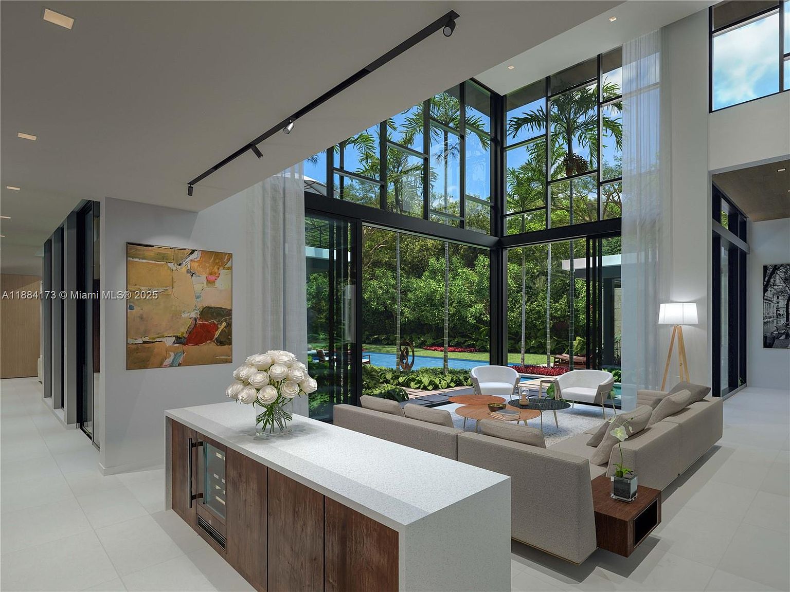 This is a spacious and modern living room with floor-to-ceiling windows offering a view of lush greenery and a pool. The room features neutral-toned sofas, a coffee table, and a stylish bar area with a wine cooler. The open floor plan and abundant natural light create an inviting and luxurious atmosphere.