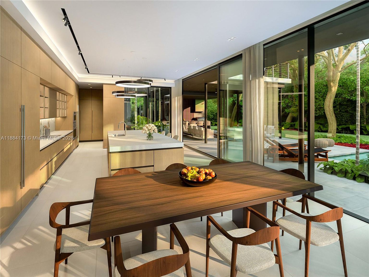 This interior shot showcases a modern dining area adjacent to a kitchen and with views of the outdoor pool area. A large wooden dining table with chairs sits prominently in the foreground, while the kitchen features sleek cabinetry and a central island. Large glass doors provide ample natural light and a seamless transition to the outdoor living space, enhancing the property's appeal.