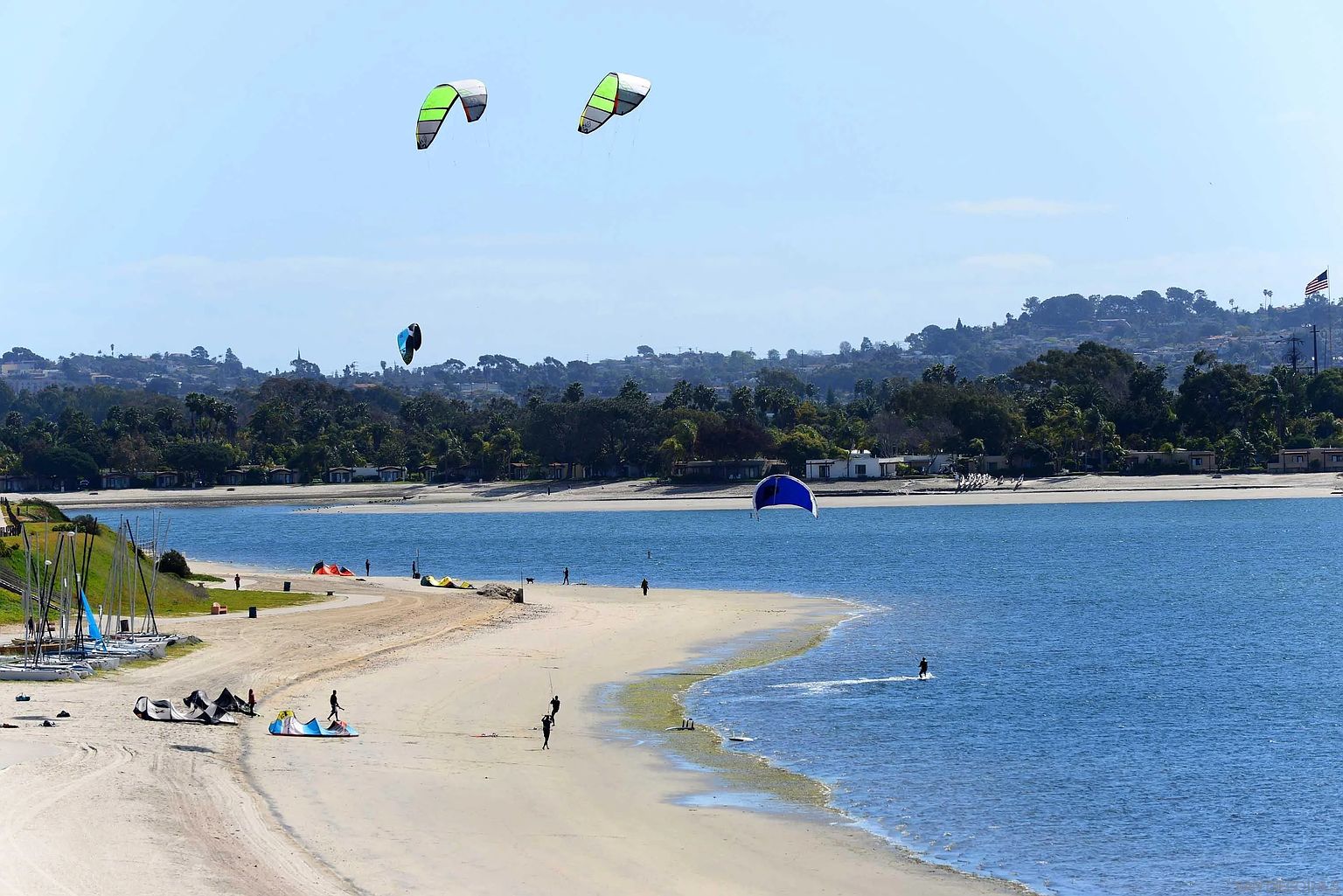 This image showcases a vibrant coastal scene, likely near a residential community. The view includes a sandy beach, calm blue waters dotted with kitesurfers, and lush green hills in the background. Several sailboats are docked along the shore, suggesting recreational opportunities are readily available.