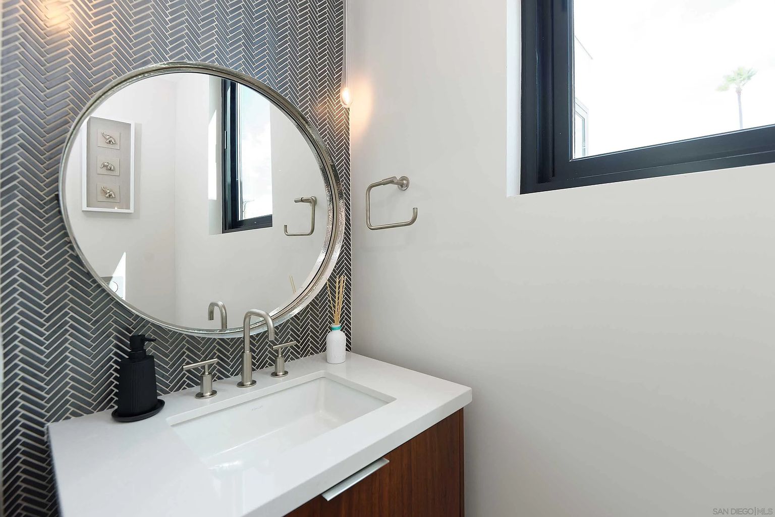 This is a well-lit guest bathroom featuring a round mirror above a modern vanity with a white countertop and wood-grain cabinetry. The wall behind the mirror is adorned with a herringbone tile pattern, adding a touch of sophistication. A black soap dispenser sits on the counter, and a window provides natural light, creating a clean and inviting space.