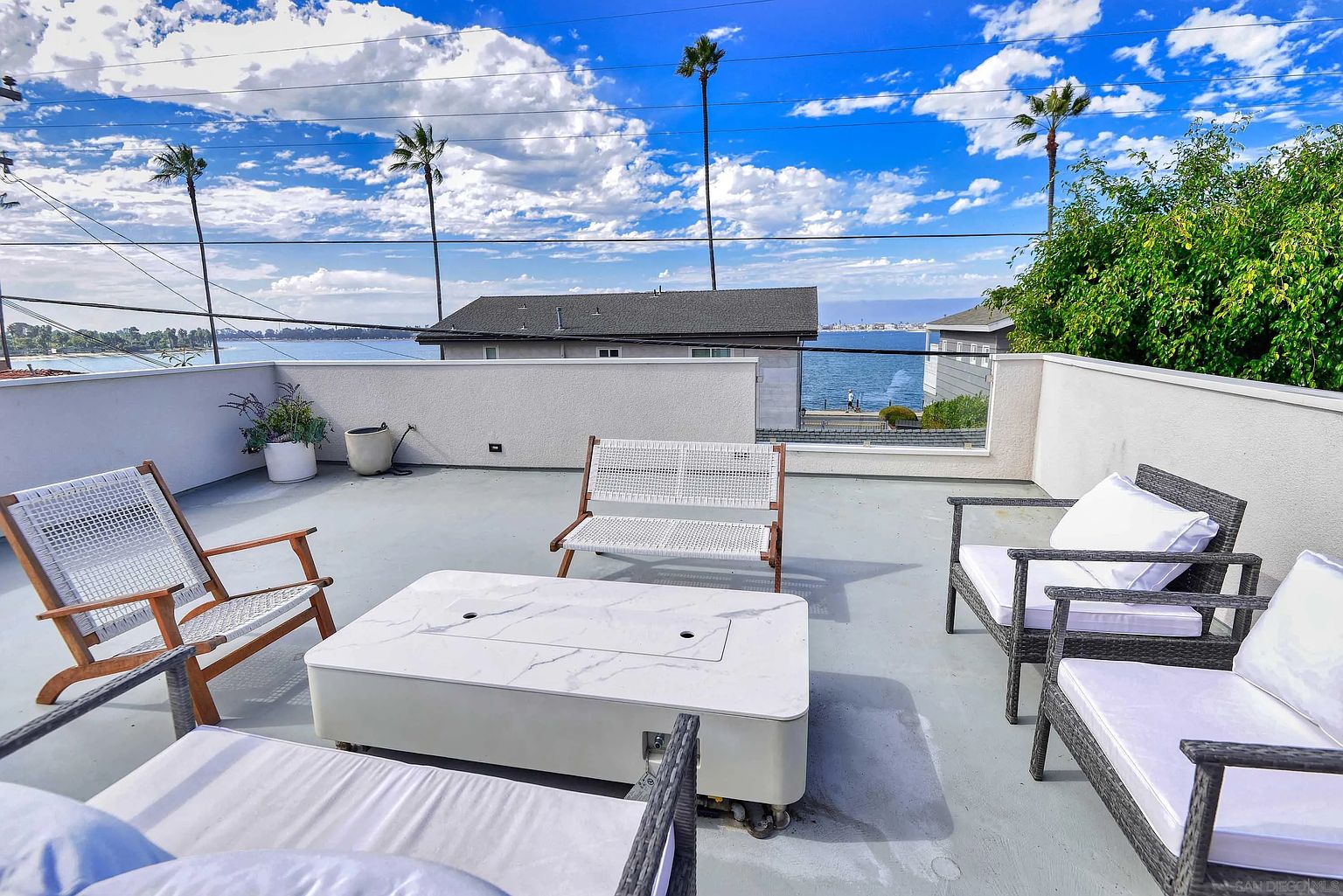 This is a stylish rooftop patio featuring modern outdoor furniture, including woven chairs and a white fire pit table. The patio offers a view of the water and surrounding landscape, with palm trees visible in the background. The space is designed for relaxation and entertainment, making it an appealing outdoor living area.