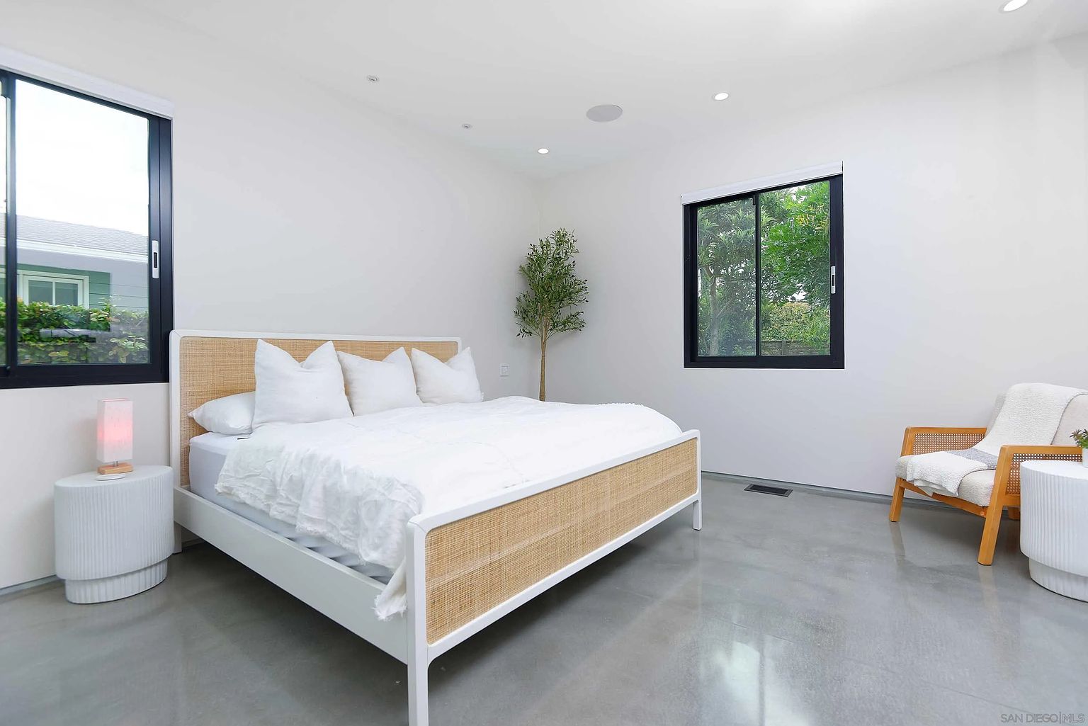 This is a bright and airy primary bedroom featuring a bed with a woven headboard and white linens. The room has polished concrete floors, white walls, and two windows that provide natural light. A small tree adds a touch of nature to the minimalist space.