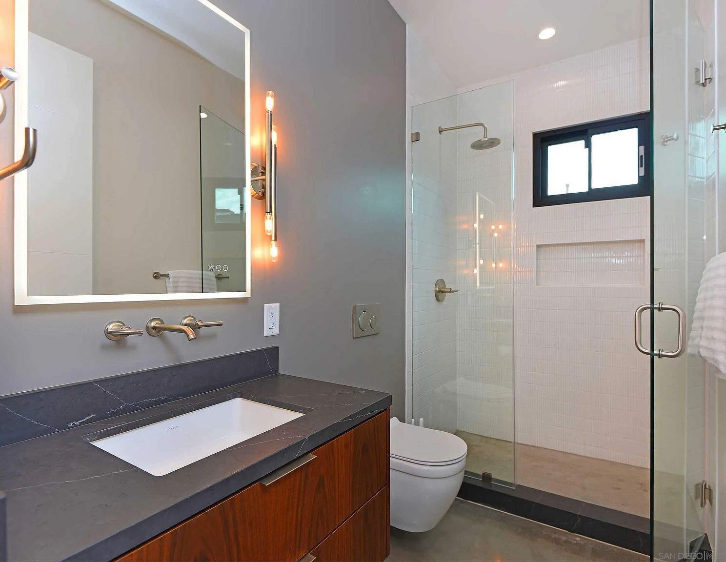 This is a modern primary bathroom featuring a sleek vanity with a dark countertop and wood cabinetry. A large, illuminated mirror hangs above the sink, complemented by stylish sconces. The walk-in shower is enclosed in glass and features white tiled walls and a black-framed window, creating a bright and contemporary space.
