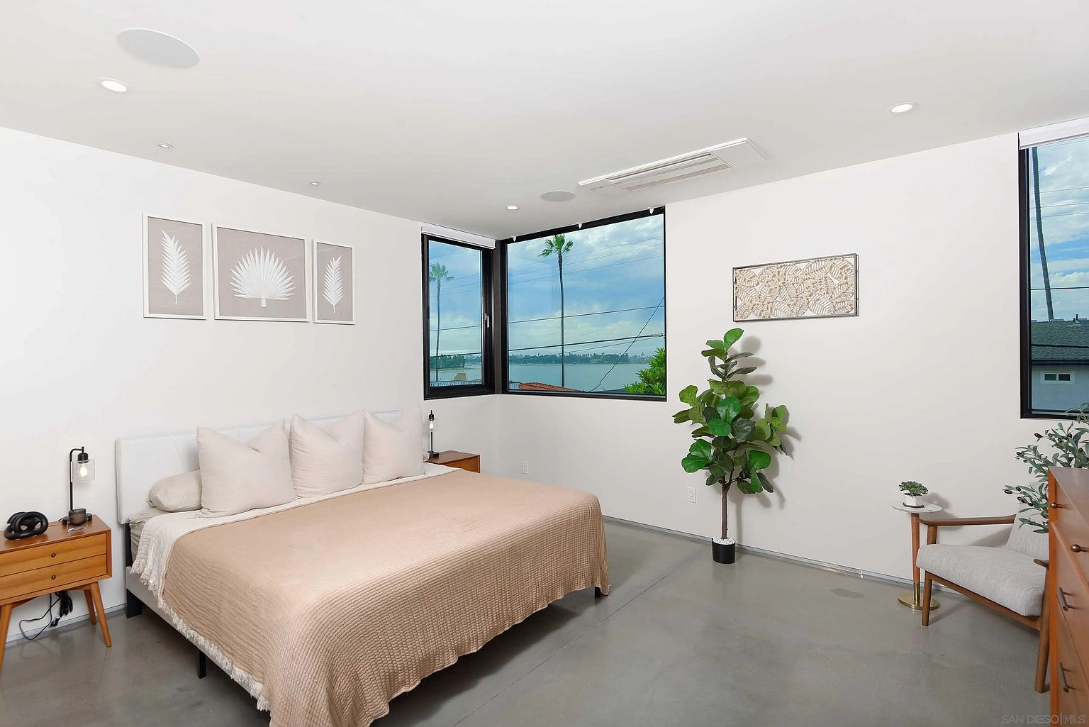 This is a bright and airy primary bedroom featuring a large bed with a light-colored quilt and pillows. The room has a modern aesthetic with white walls, polished concrete floors, and large windows offering a view of the water. The room is decorated with minimalist artwork and a potted fiddle-leaf fig tree, creating a serene and inviting atmosphere.