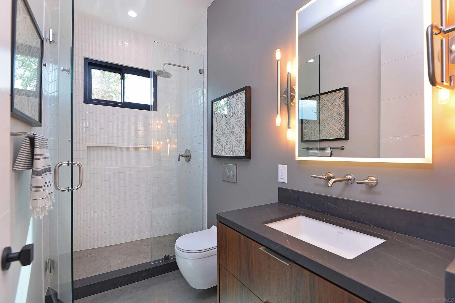 This is a modern bathroom featuring a glass-enclosed shower with white vertical tiles and a black-framed window. The vanity has a dark countertop and wood-grain cabinets, complemented by a backlit mirror and stylish sconces. The overall design is sleek and contemporary, creating a spa-like atmosphere.