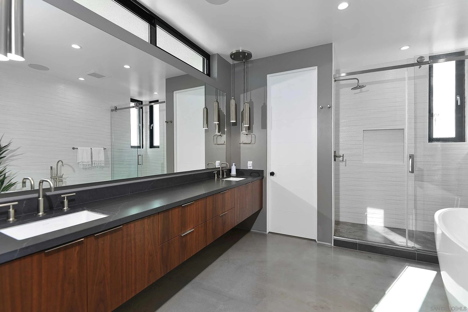 This is a modern primary bathroom featuring a long, floating vanity with dark countertops and wood-grain cabinets. A large mirror reflects the space, including a glass-enclosed shower with white tiled walls and a separate white door. The flooring is a sleek gray, and pendant lights hang above the vanity, adding a touch of elegance.