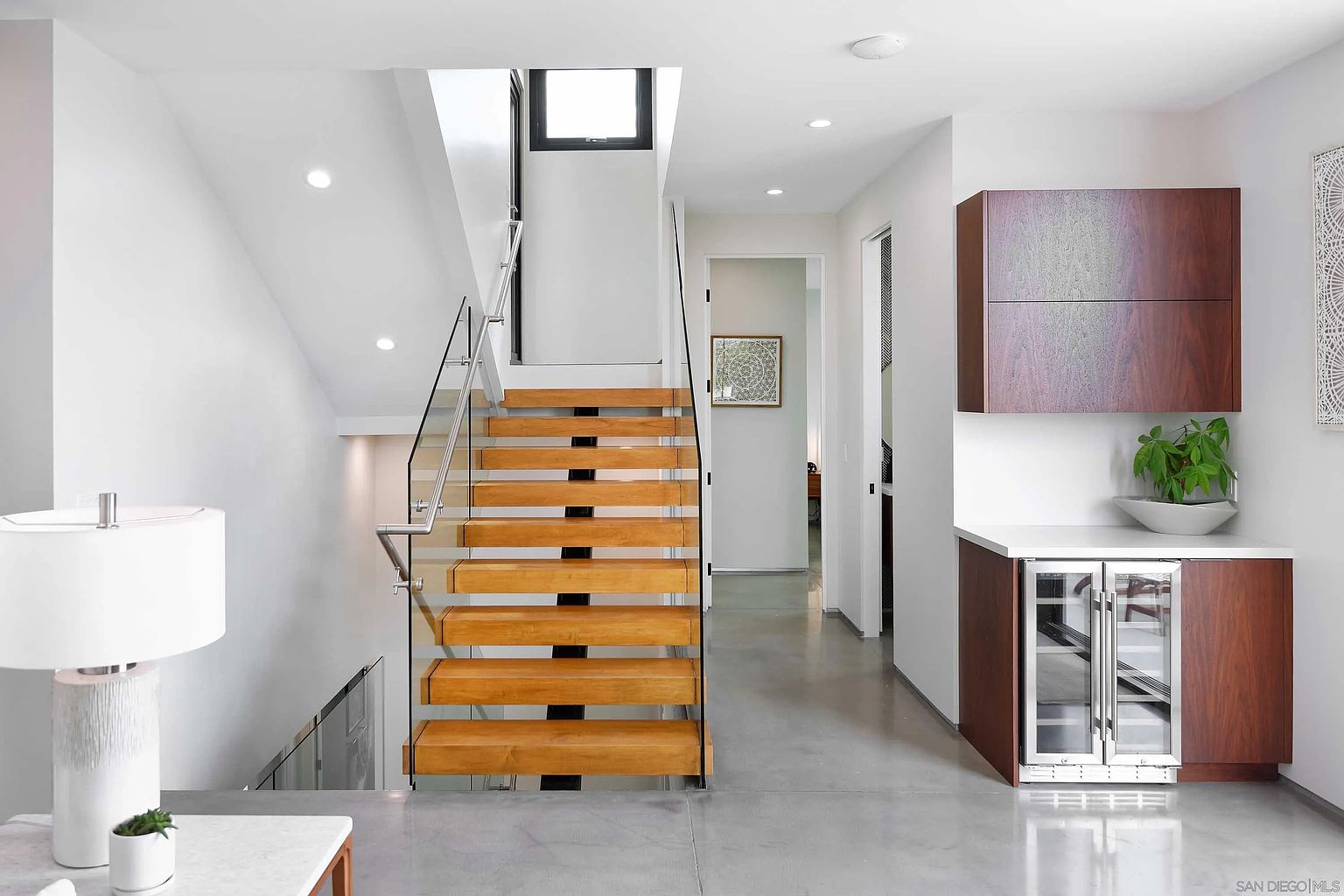 This interior shot showcases a modern hallway and staircase. The staircase features open wooden treads with glass railings, leading to an upper level. A built-in bar area with a wine fridge adds a touch of luxury, while the polished concrete floors contribute to the contemporary aesthetic. The space is well-lit and appears clean and minimalist.