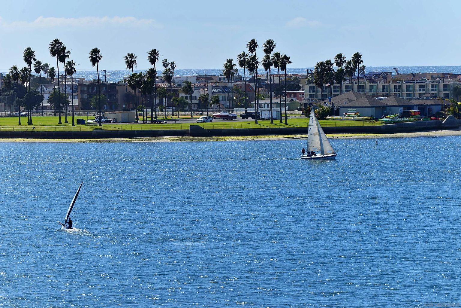 This image showcases a scenic waterfront view with palm trees lining the shore, suggesting a coastal community amenity. A sailboat and windsurfer are visible on the water, indicating recreational opportunities. The presence of buildings in the background suggests residential development near the water.