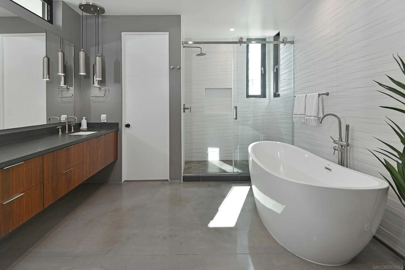 This is a modern primary bathroom featuring a freestanding white bathtub, a glass-enclosed shower, and a floating vanity with a dark countertop and wood-grain cabinets. The walls are a mix of gray paint and textured white tile, and the floor is polished concrete. The overall impression is clean, minimalist, and luxurious.