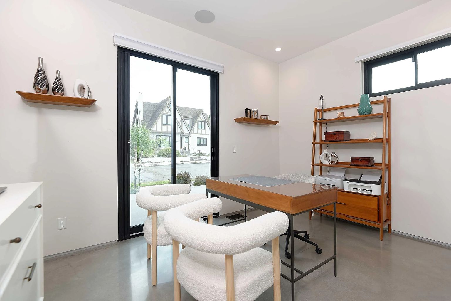 This is an interior shot of a modern home office. The room features a wooden desk with a gray inset, two white boucle chairs, and a wooden shelving unit. A large sliding glass door provides natural light and a view of the exterior, while a smaller window is visible on the right wall. The overall aesthetic is clean, minimalist, and inviting, perfect for a productive workspace.