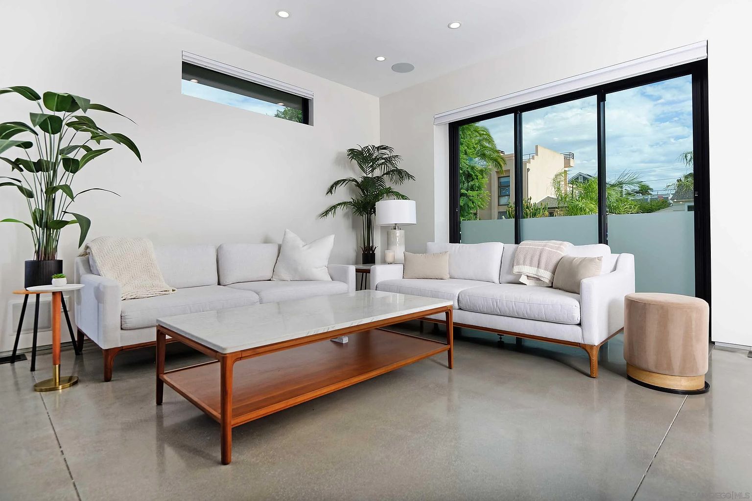 This is a bright and airy living room featuring two white sofas, a marble-topped coffee table with a wooden base, and large windows that let in plenty of natural light. The room is decorated with plants, adding a touch of nature to the modern space. The polished concrete floors and minimalist decor contribute to a clean and contemporary aesthetic.