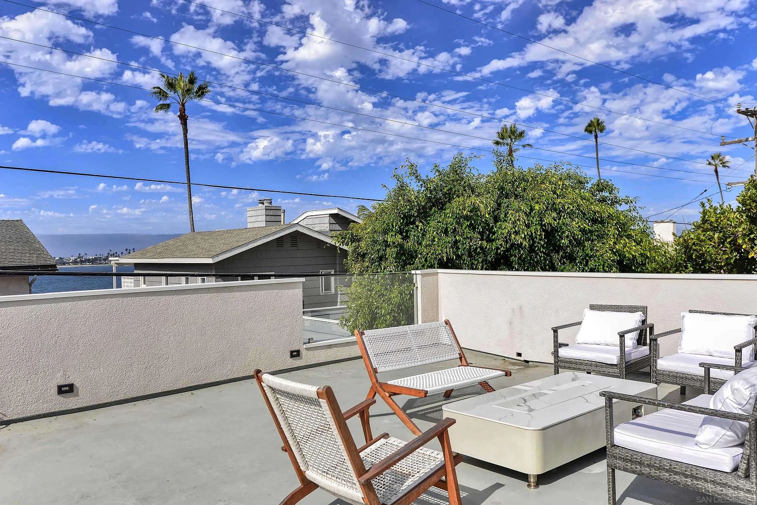 This image showcases a stylish rooftop patio or deck area, featuring modern outdoor furniture including woven chairs and a sleek fire pit table. The space is enclosed by a low wall and glass railing, offering views of the surrounding neighborhood, including palm trees and glimpses of the ocean. The setting suggests a relaxed and upscale outdoor living space, perfect for entertaining or enjoying the scenery.