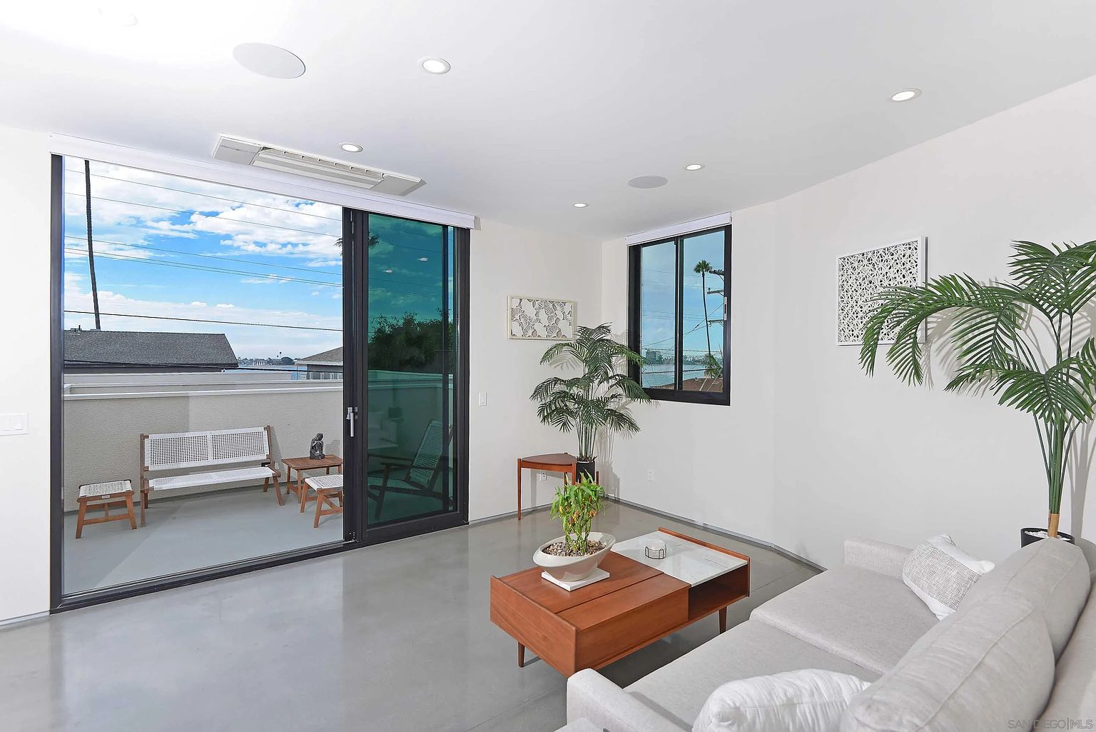 This is a bright and airy living room featuring a modern design with clean lines and a neutral color palette. The room has a large sliding glass door leading to a balcony, providing ample natural light and a view of the outdoors. The space is furnished with a comfortable sofa, a stylish coffee table, and several potted plants, creating a welcoming and relaxing atmosphere.