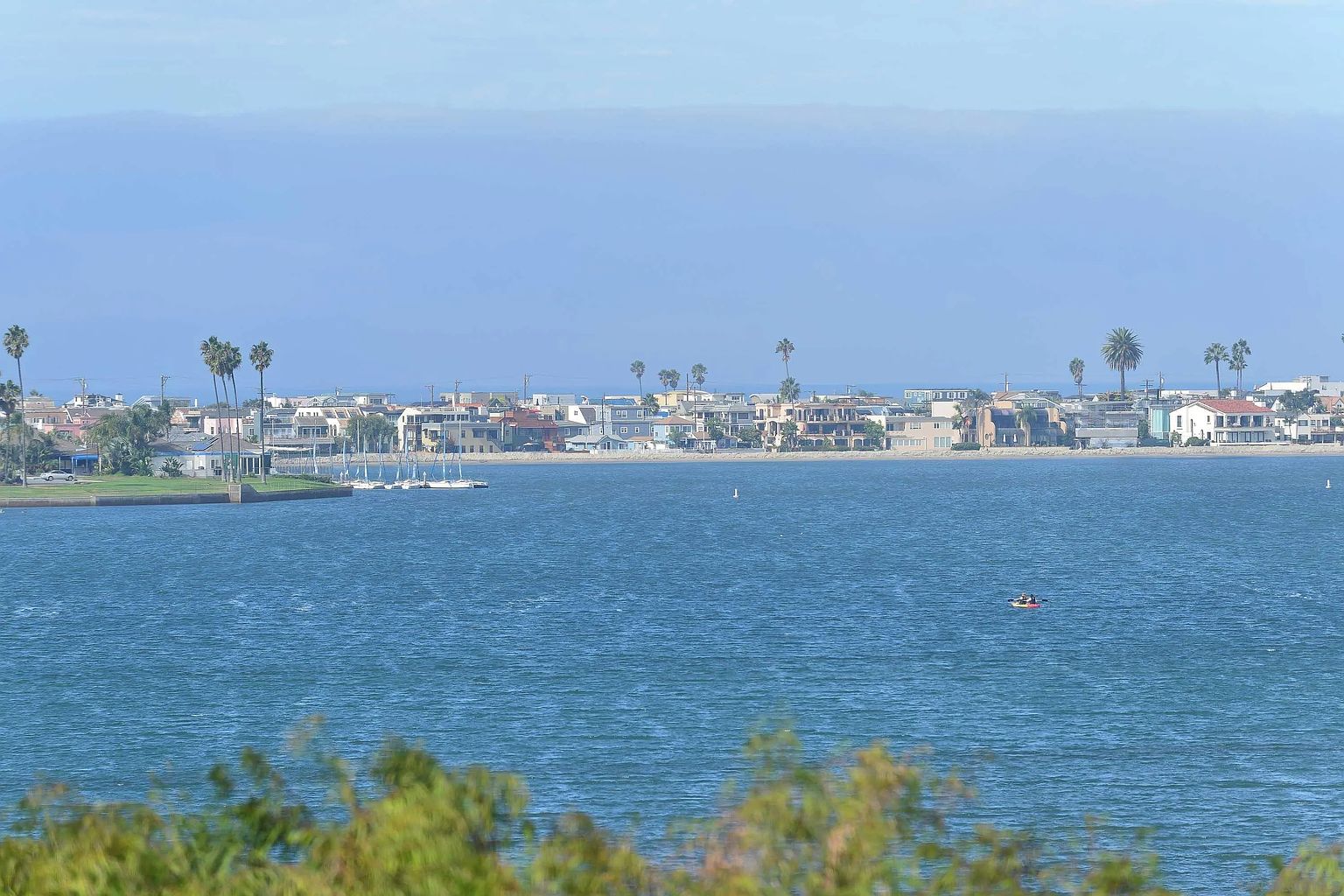 This aerial view showcases a waterfront community with houses lining the coast, palm trees dotting the landscape, and boats gently sailing on the blue water. The scene evokes a sense of tranquility and coastal living, highlighting the desirable location and lifestyle offered by these properties. The perspective is a wide shot, capturing the expansive water and the community nestled along the shore.