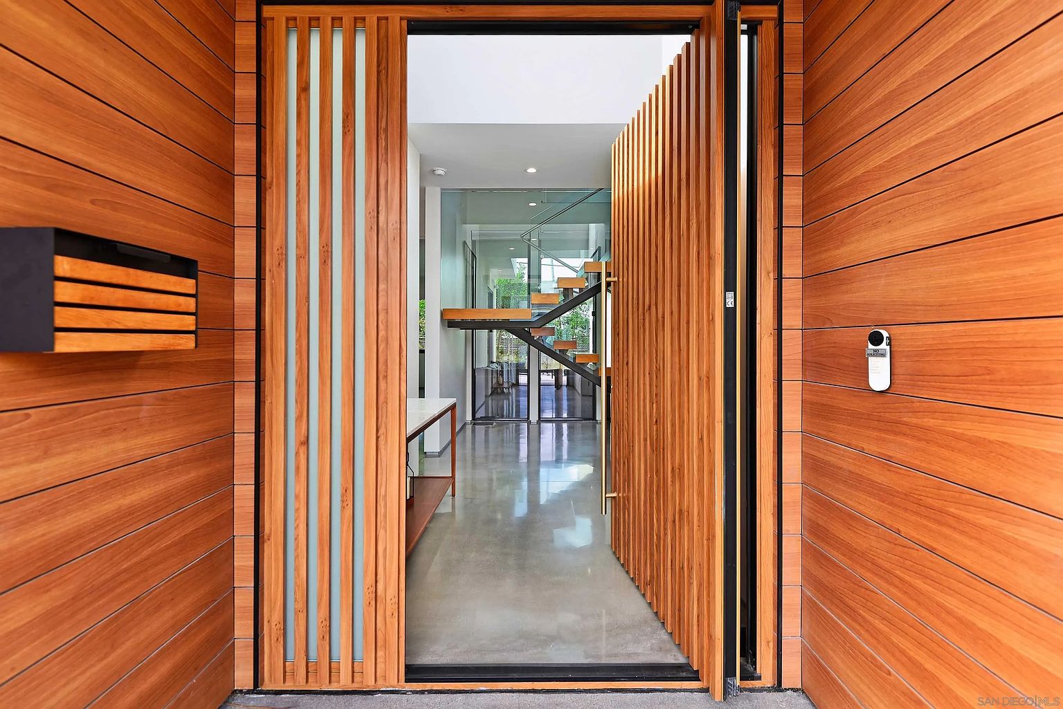 This image showcases a modern home's entryway, featuring a striking wooden door with vertical slats and glass panels. The exterior walls are clad in horizontal wood planks, creating a warm and inviting aesthetic. The open door reveals a glimpse of the interior, including a staircase and polished concrete floors, suggesting a contemporary and stylish living space.